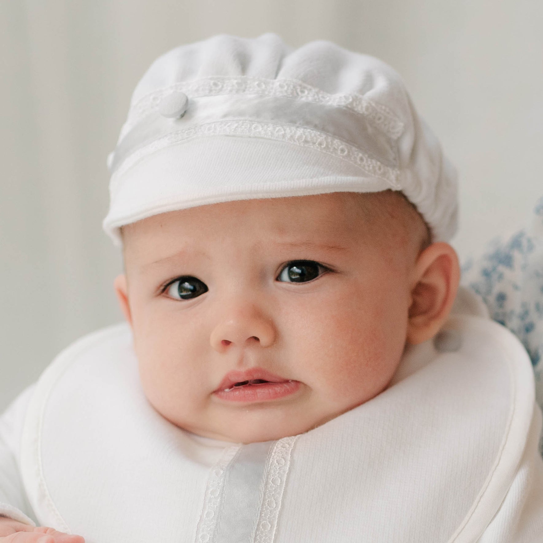Baby in the Henry Brim Cap and matching 3-Piece Suit and Bib sitting on a woman with blue patterned dress. Part of Christening Gowns' high end baby clothes.