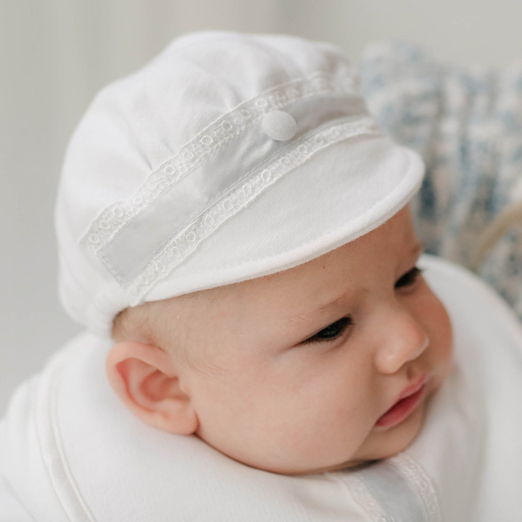 Close up of a baby wearing the Henry Brim Cap with lace details on a neutral background. The hat is crafted with white french terry and features Venice lace and blue silk ribbon with a blue linen button.