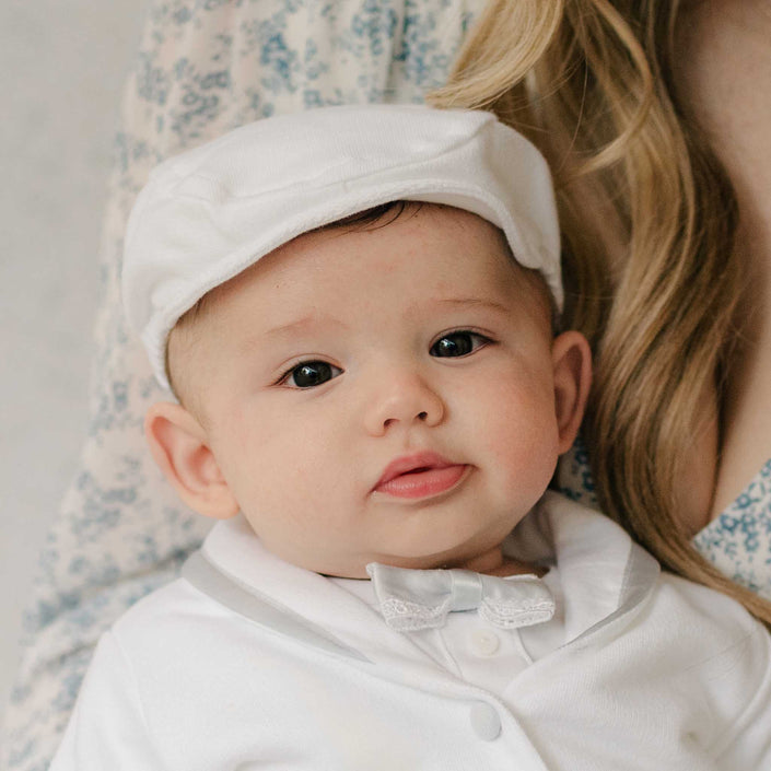 Close up of a baby in the Henry Newsboy Cap and matching 3-Piece Suit being held by a woman in a floral-patterned fabric dress. Details show the cap crafted with white French Terry cotton. Part of Baby Beau & Belle's luxury baby clothes.