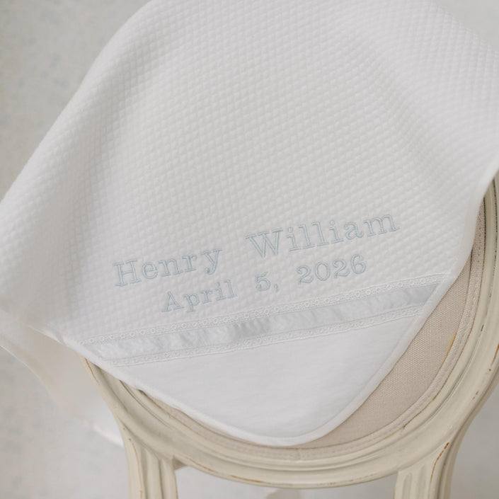 Close up photo of the Henry Personalized Blanket draped on a beige chair. The quilt blanket has the name "Henry" embroidered on the corner. Perfect baptism gift ideas.