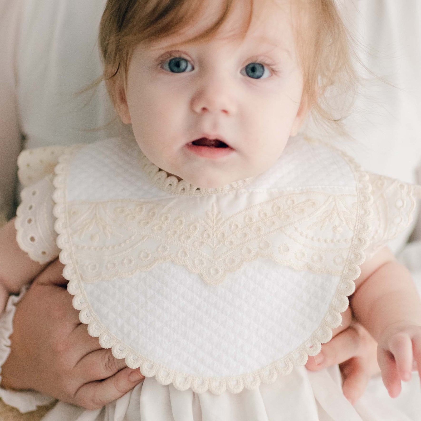 Baby wearing the Ingrid Bib crafted with a soft quilted cotton, accented with a sweet scalloped ecru edge lace and detailed with the matching Ingrid eyelet lace