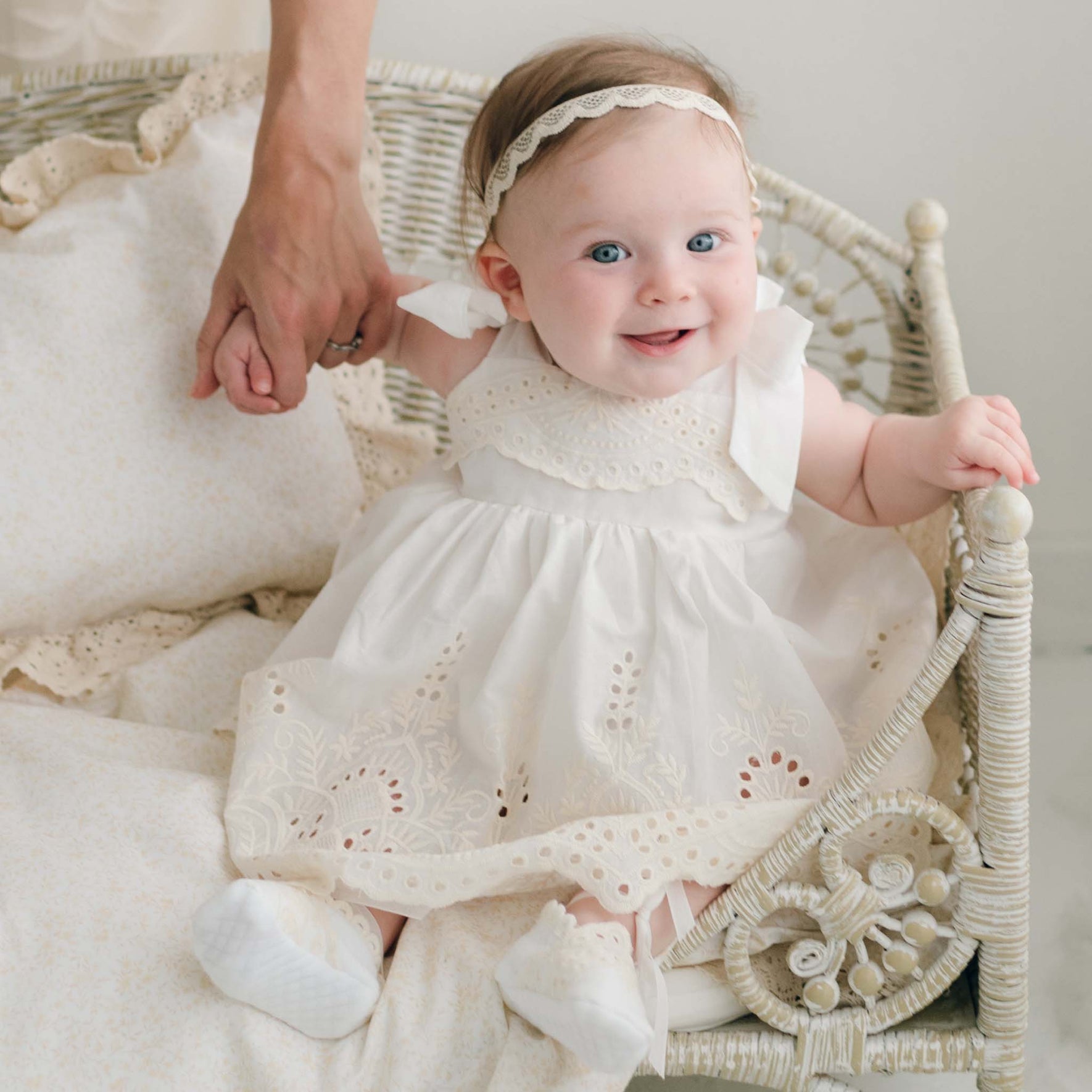 An infant sits smiling in a wicker crib, dressed in the Ingrid Romper Dress made from 100% cotton with delicate lace detailing. The baby holds an adult's hand and wears a matching headband and white socks, creating a charming contrast against the light-colored pillows in the background.
