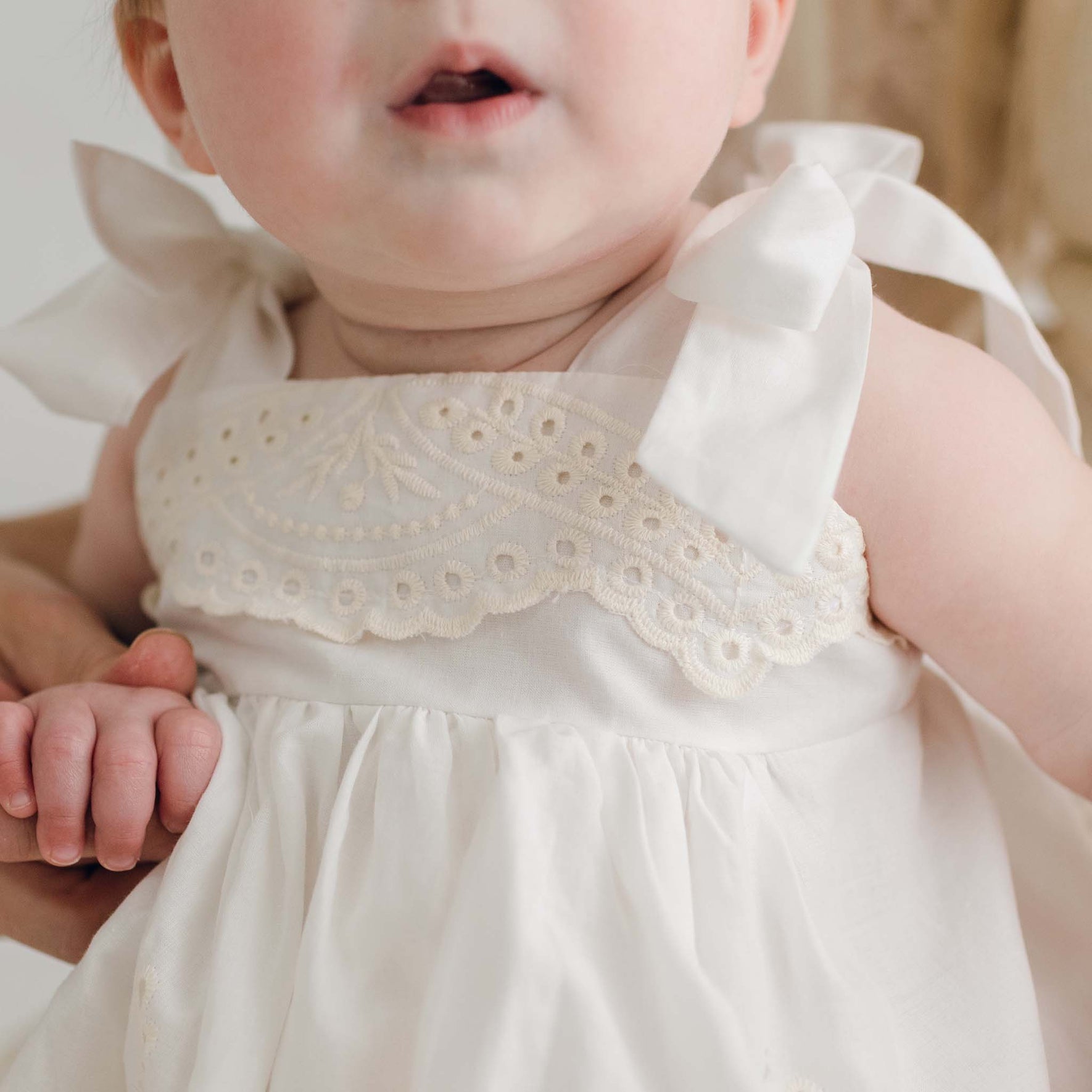 A close-up of a baby wearing an Ingrid Romper Dress, which features intricate lace details at the top and delicate ribbon straps. An adult is gently holding the baby's hand, while the lower half of the baby's face is in focus.