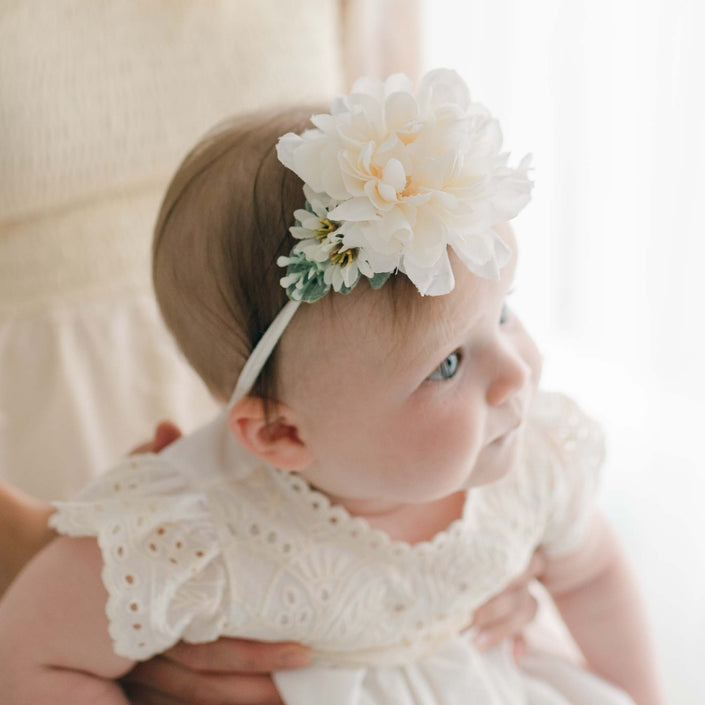 An infant adorned in a white dress and the Ingrid Flower Headband, featuring an ivory dahlia, is cradled by an unseen person. The super soft nylon band adds a touch of comfort as the baby gazes slightly to the right.