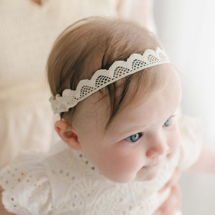 A baby wearing an Ingrid Lace Headband and a matching white lace outfit, reminiscent of an Ingrid Christening Gown, is held by an adult whose face is not visible. The baby has light hair and blue eyes and is looking slightly to the side.