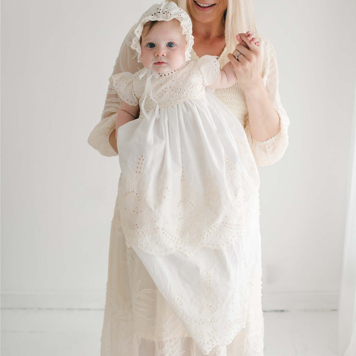 A woman, whose face is not visible, holds a baby dressed in the Ingrid Tiered Gown & Bonnet—a handmade in the USA white lace christening ensemble. The minimalist aesthetic is enhanced by the plain white background as the baby looks directly at the camera.
