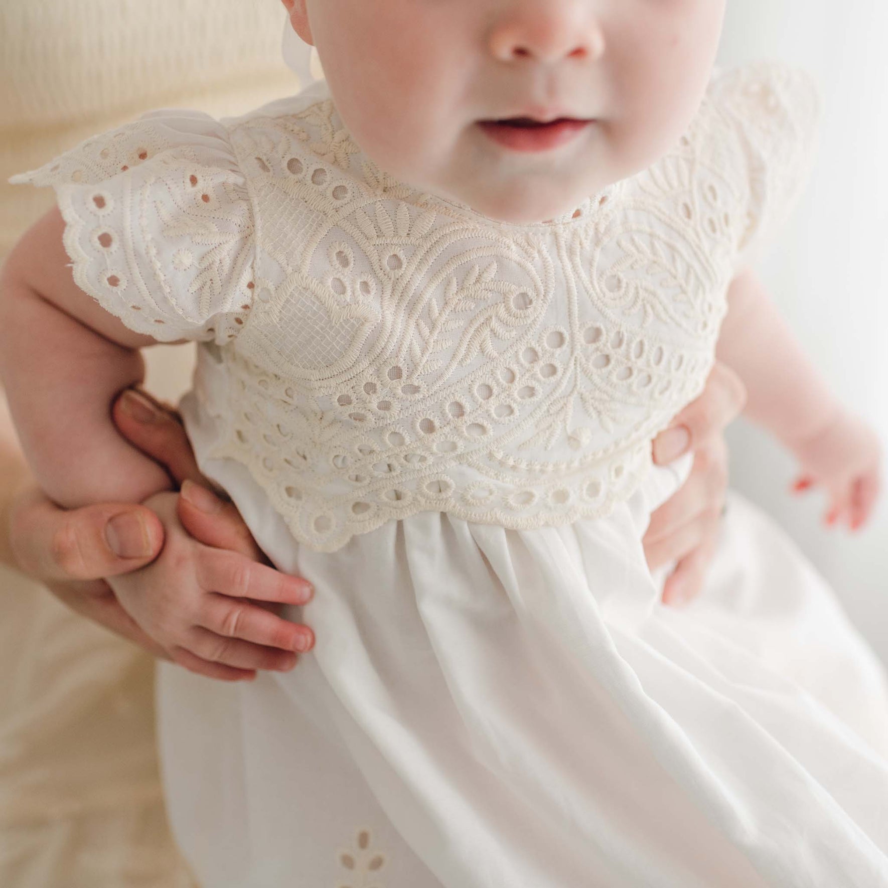 Close-up of a baby in an Ingrid Tiered Gown & Bonnet, gently held by an adult's hands. The Christening gown features intricate lace detailing on the bodice and sleeves, with small eyelets and embroidered patterns. The baby's face is partially visible, showing their lips and nose.