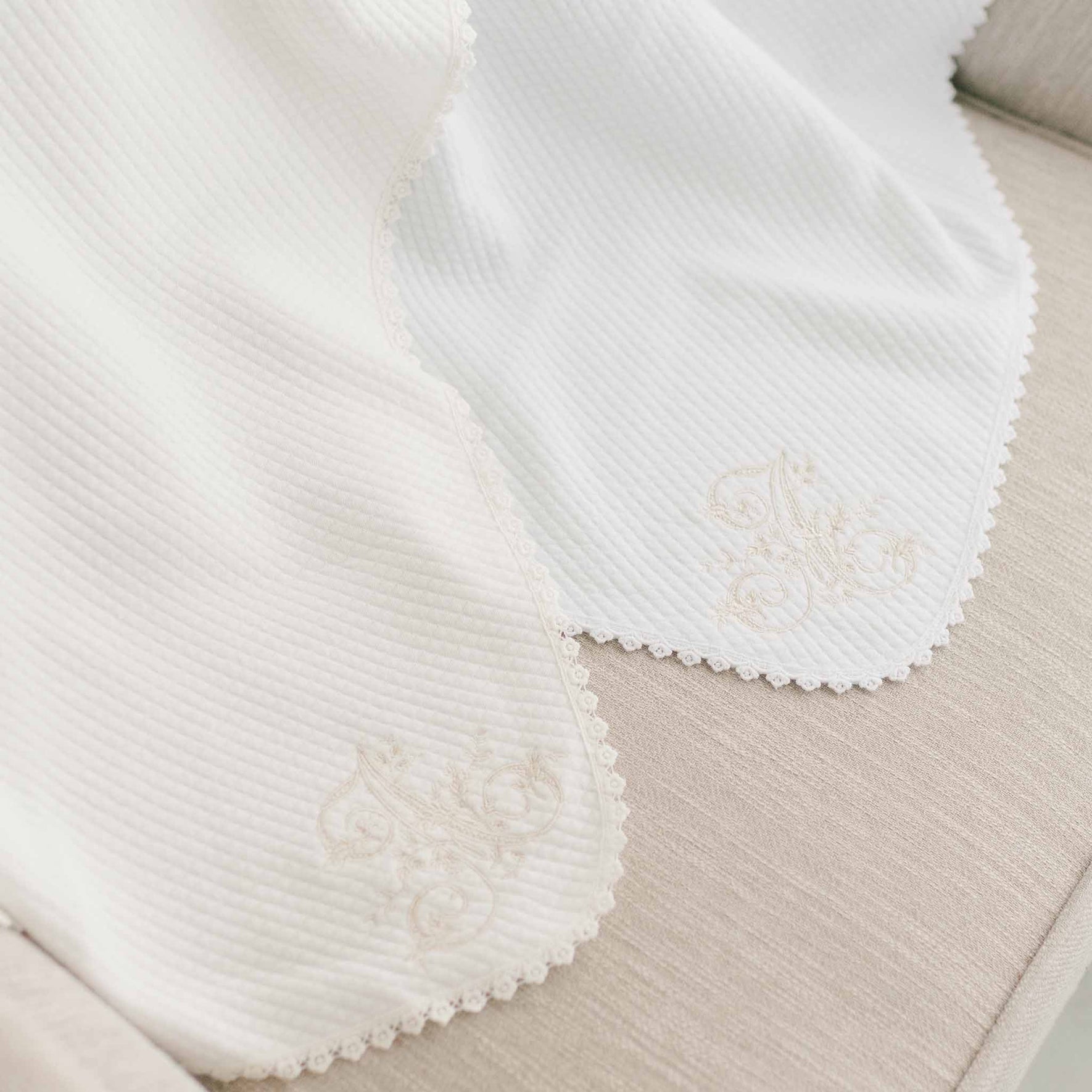 A close-up of the Initial Blanket reveals its light beige quilted cotton fabric, featuring delicate floral embroidery with personalized initials along the edge and bordered by intricate lace trim. The blanket is elegantly draped over a matching surface.