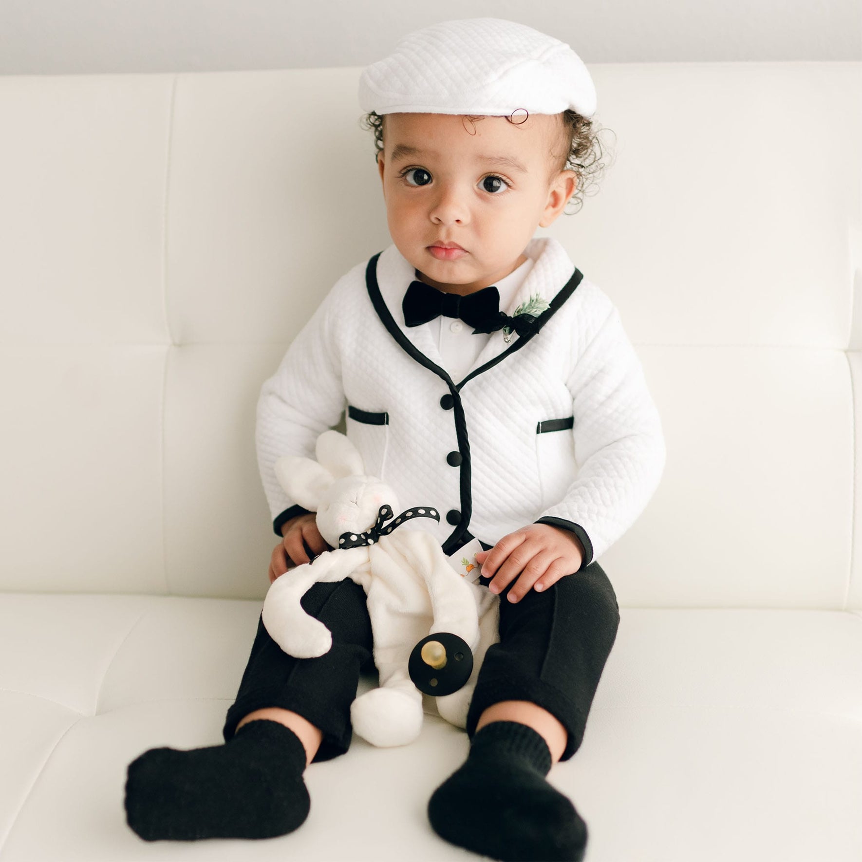 A baby sitting on a white couch in the James 3-Piece Suit, holding a plush bunny, with black pants and socks complementing the outfit.