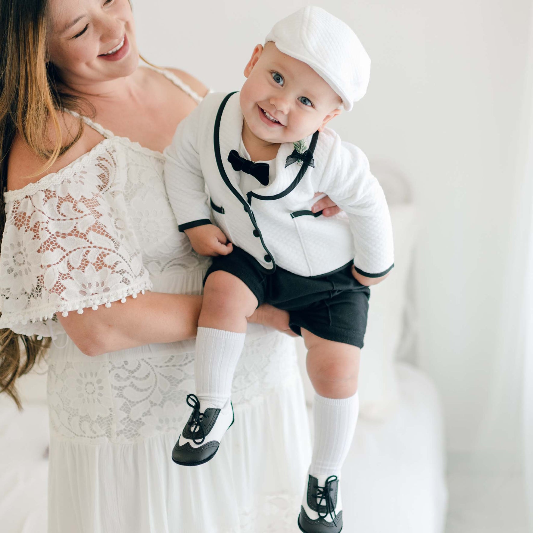 A baby dressed in the James 3-Piece Suit, featuring a quilted white jacket with black trim, black shorts, and a matching white cap, held in a mother's arms.