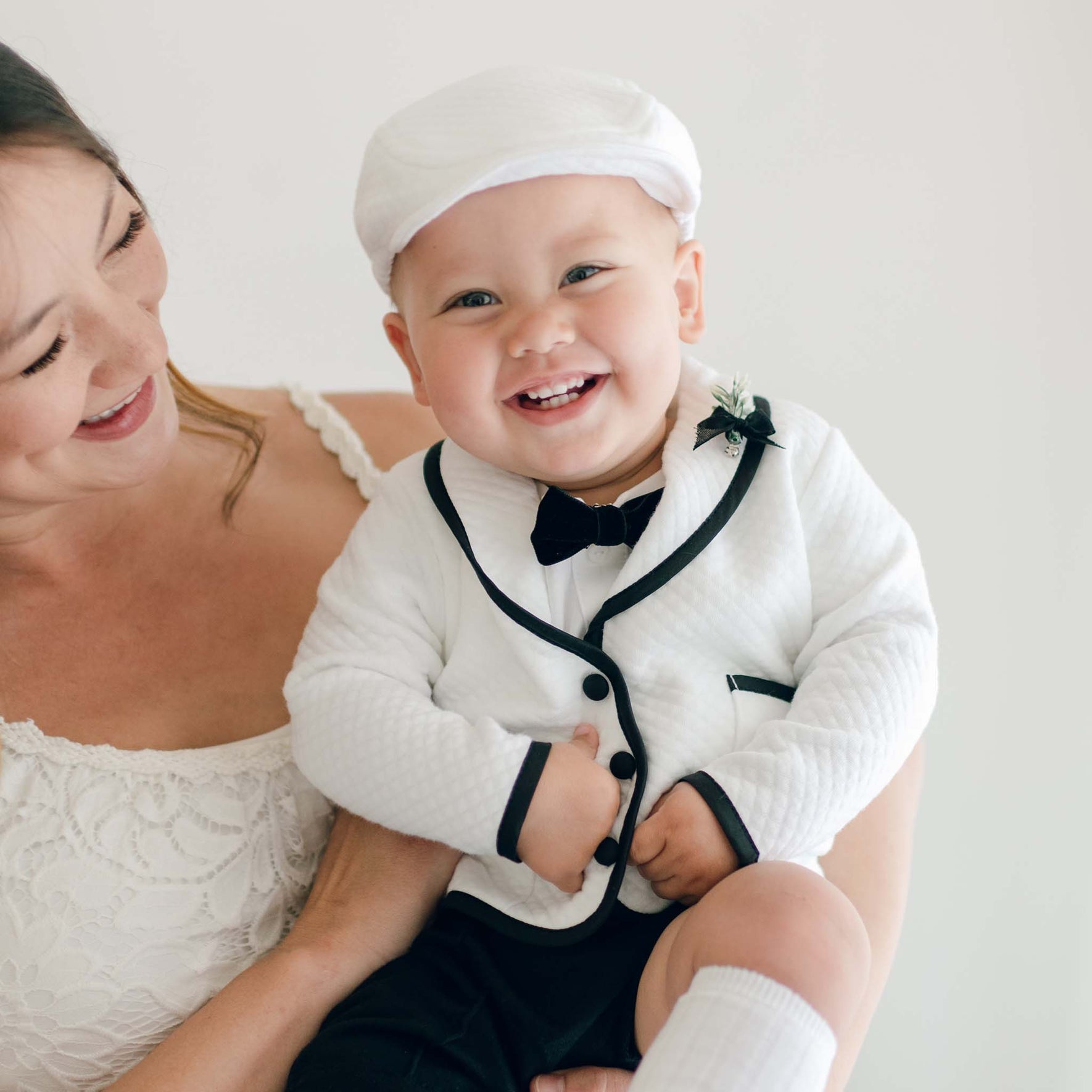 A baby wearing the James 3-Piece Suit sits on a parent's lap, showcasing the white jacket with black trim, black bow tie, and decorative boutonniere.
