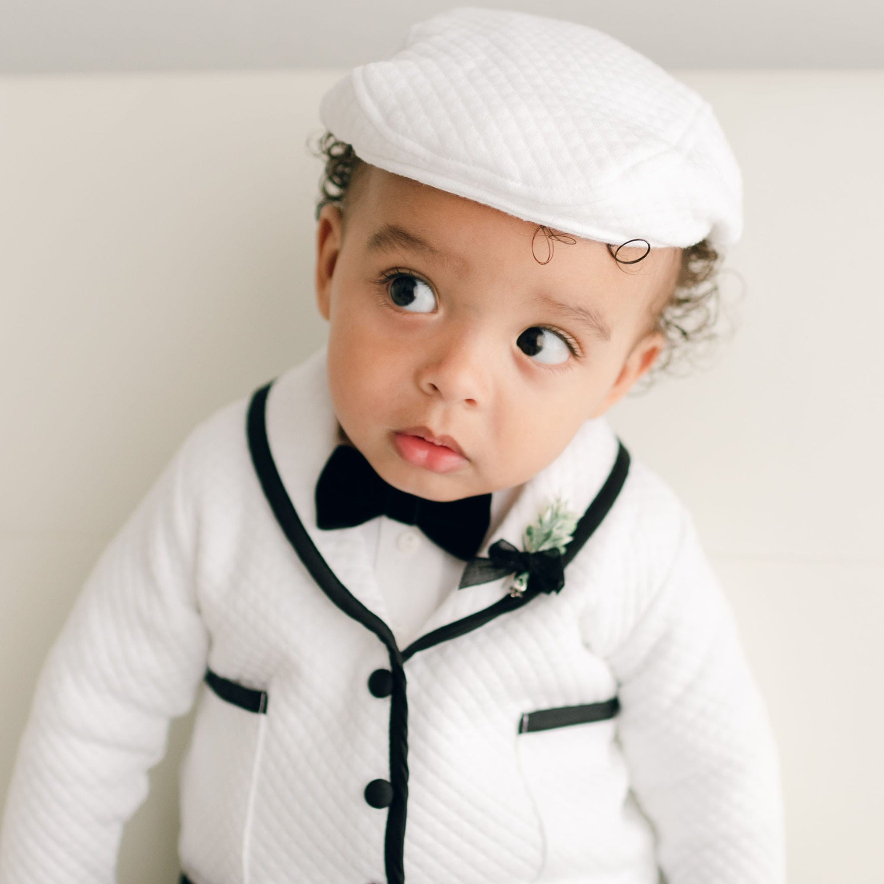 A close-up of a baby in the James 3-Piece Suit, highlighting the white quilted jacket, black bow tie, and matching cap.