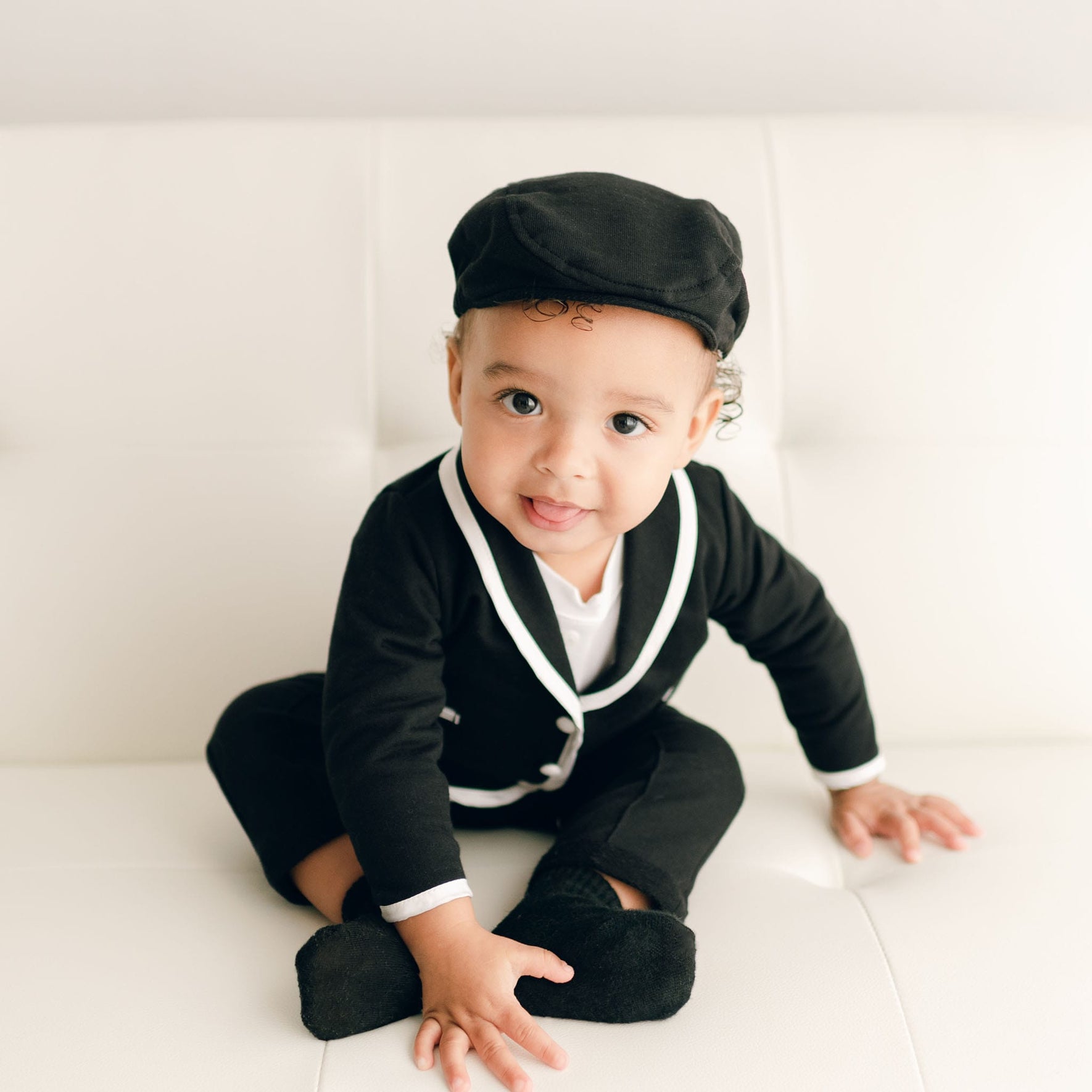 A baby boy sitting on a white couch in the James 3-Piece Suit in black, playfully sticking out his tongue, highlighting the outfit’s tailored fit.
