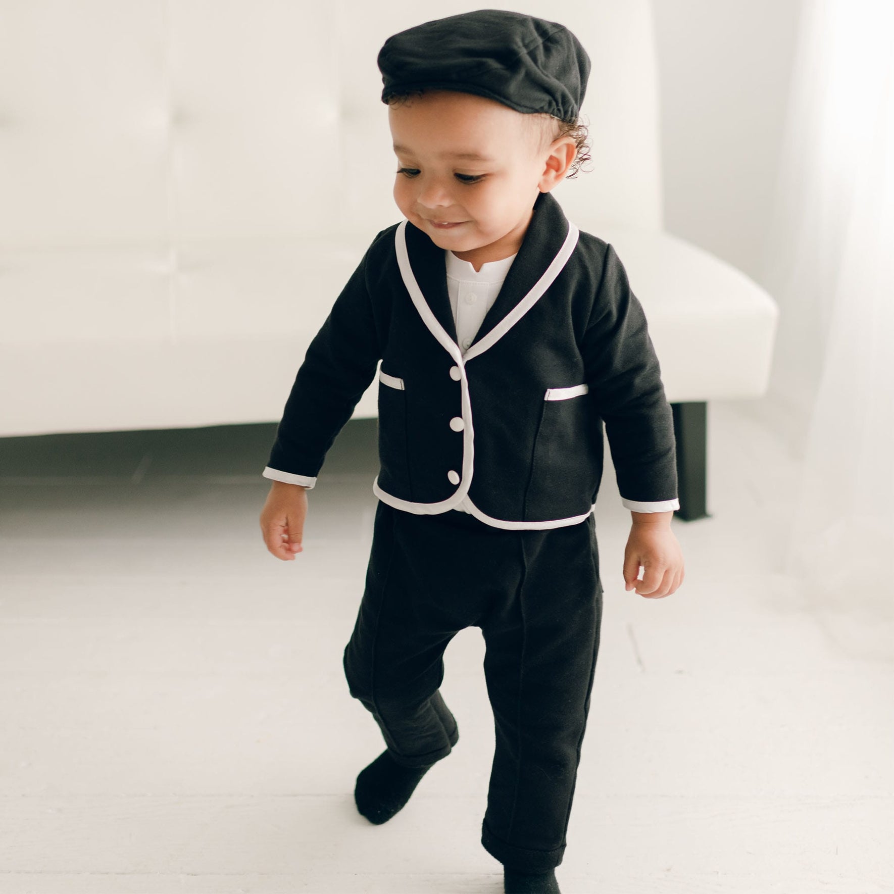 A baby boy in the James 3-Piece Suit in black, smiling while taking steps, wearing a black cap and a white shirt underneath the jacket.
