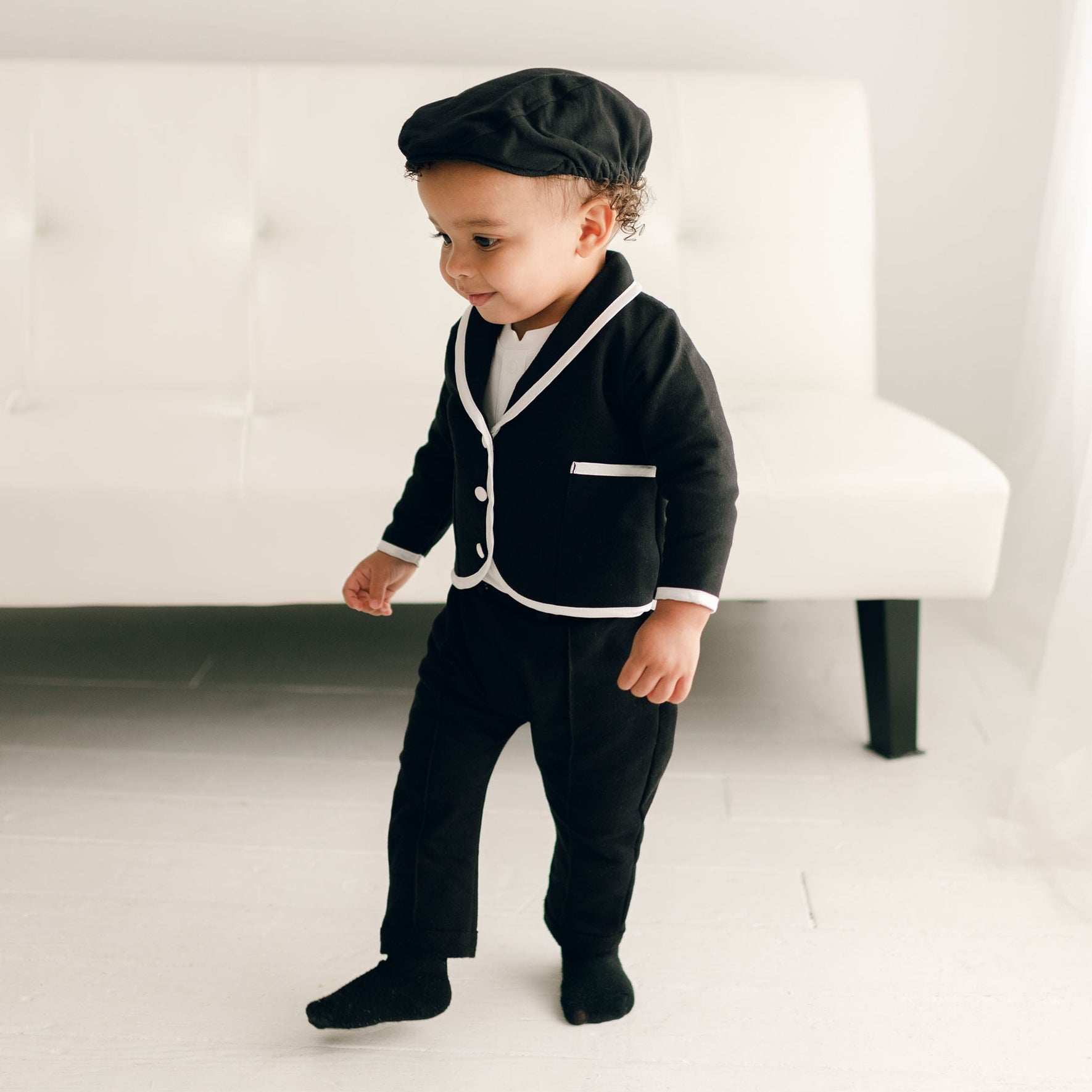A baby boy wearing the James 3-Piece Suit in black, walking near a white couch, showcasing the black jacket with white trim and matching pants.