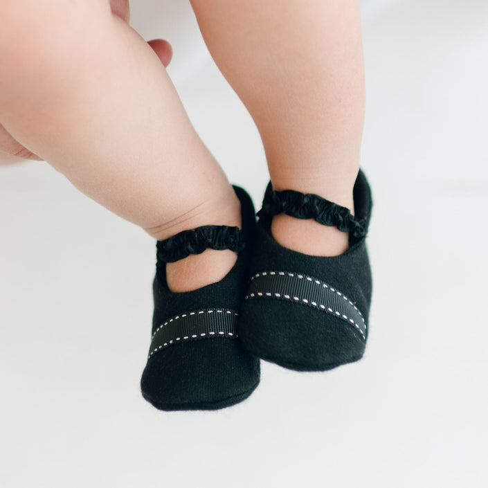 Close-up of a baby's legs wearing black James Booties featuring elastic ankle straps and detailed white stitching. The background is white and out of focus.