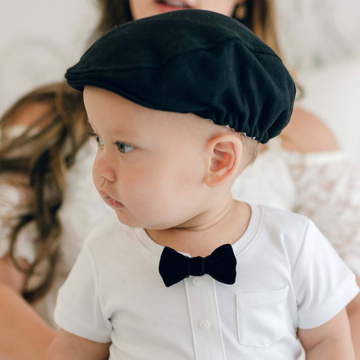 A baby, looking to the side, wears a white shirt with buttons, a black bow tie, and a stylish James Newsboy Cap. The baby is also dressed in an elegant christening outfit. An adult, whose face is partially visible, stands in the background.