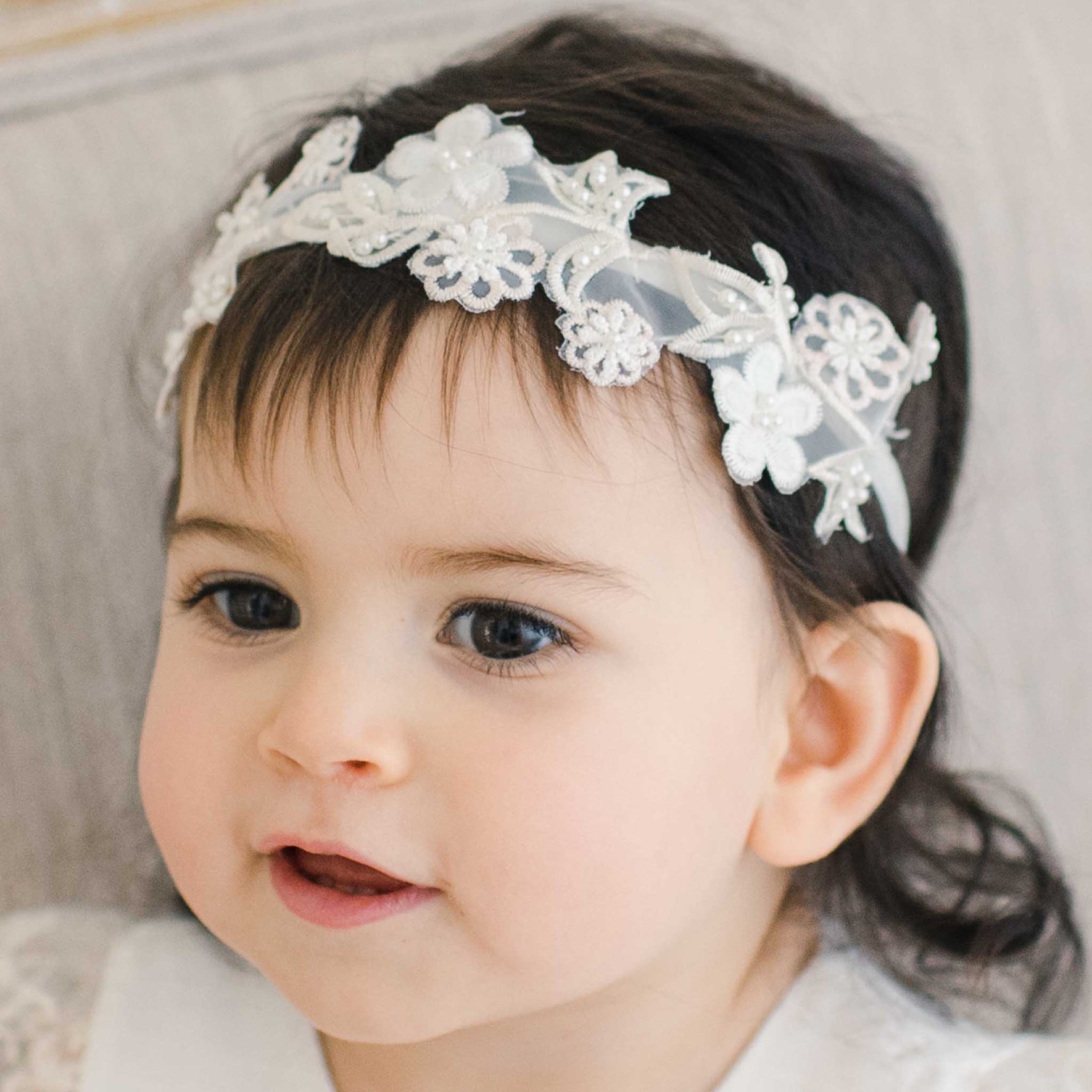 A young child with dark hair, wearing a Jessica Beaded Flower Headband, looks slightly to the side. They have a soft expression on their face. The background is a neutral, light-colored surface.