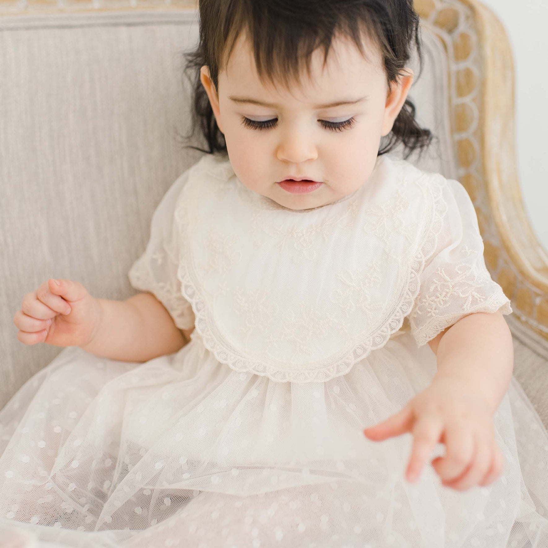 A young child with dark hair is sitting on a light-colored upholstered chair. The child is wearing a light-colored, lacy dress and a delicate Jessica Cotton Christening Bib. The child is looking down and touching the fabric of the dress with both hands.