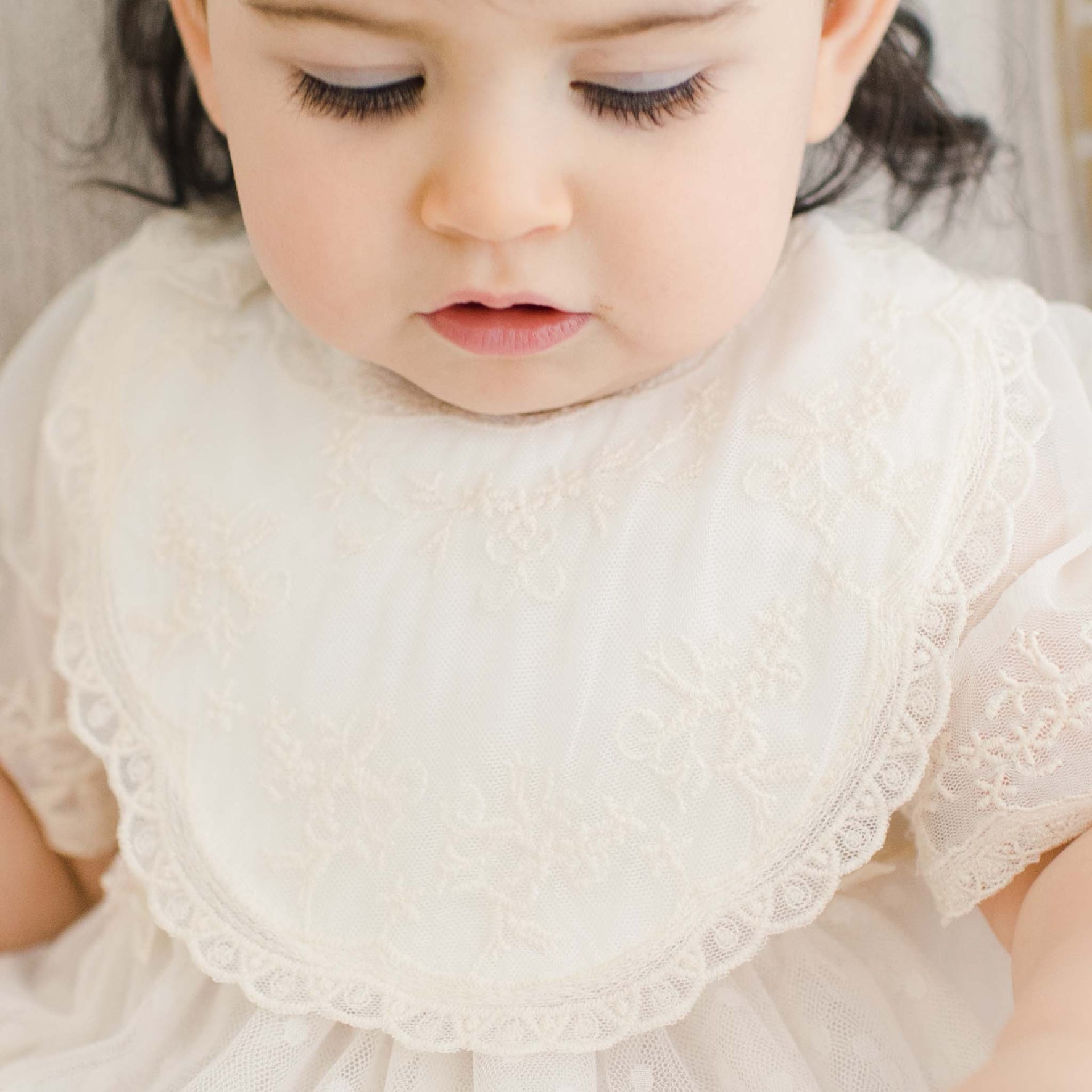 A close-up of a young child wearing a white lace dress with floral embroidery on the Jessica Cotton Christening Bib. The child has dark hair and is looking downward, with their eyelids partially closed. Their lips are slightly parted.