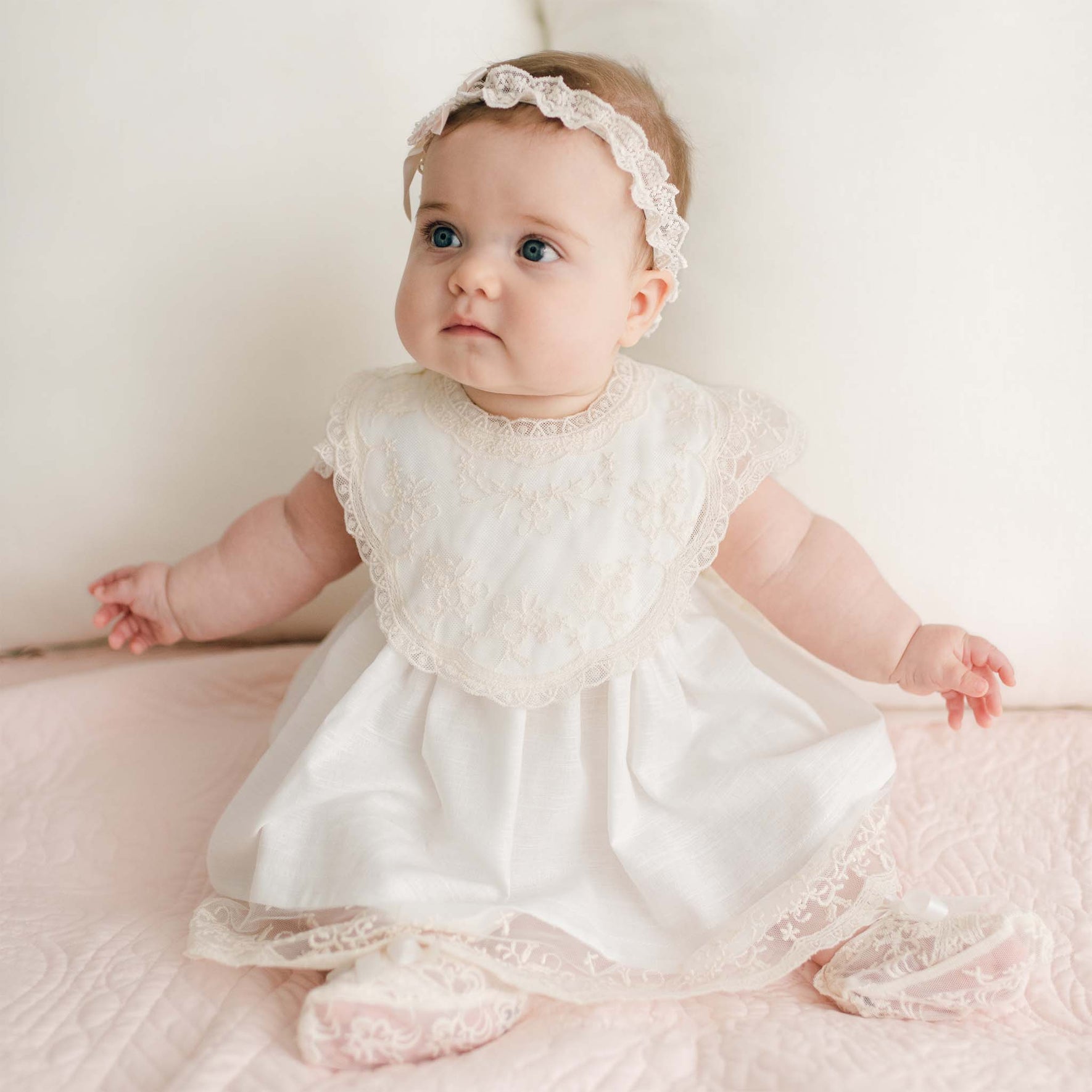 A baby wearing a white champagne lace dress and headband sits on a pink quilted surface. The baby, with a neutral expression, looks slightly to the right. Beside the baby, a Jessica Cotton Christening Bib adds charm to the scene. The background consists of white cushions, creating a serene atmosphere.