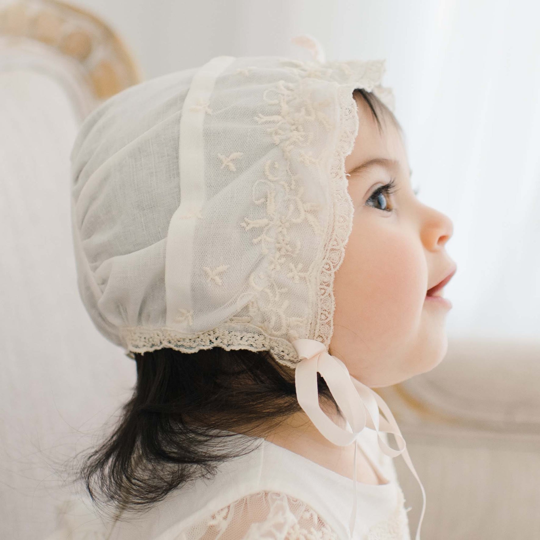 A young child is pictured in profile wearing a white Jessica Linen Bonnet with Champagne Lace details. The child's dark hair is partially visible under the bonnet, and an ivory ribbon ties under the chin. The background is softly blurred, focusing attention on the child's face and attire.