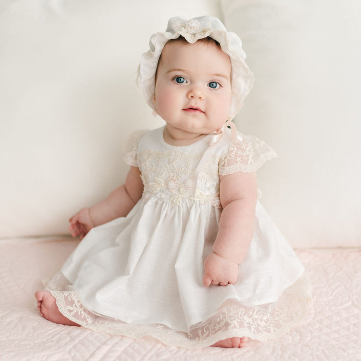 A baby is sitting on a soft surface, dressed in the Jessica Linen Dress & Bloomers along with a matching bonnet. The background showcases light-colored quilted fabric, lending a vintage touch. The baby gazes upward with a neutral expression.