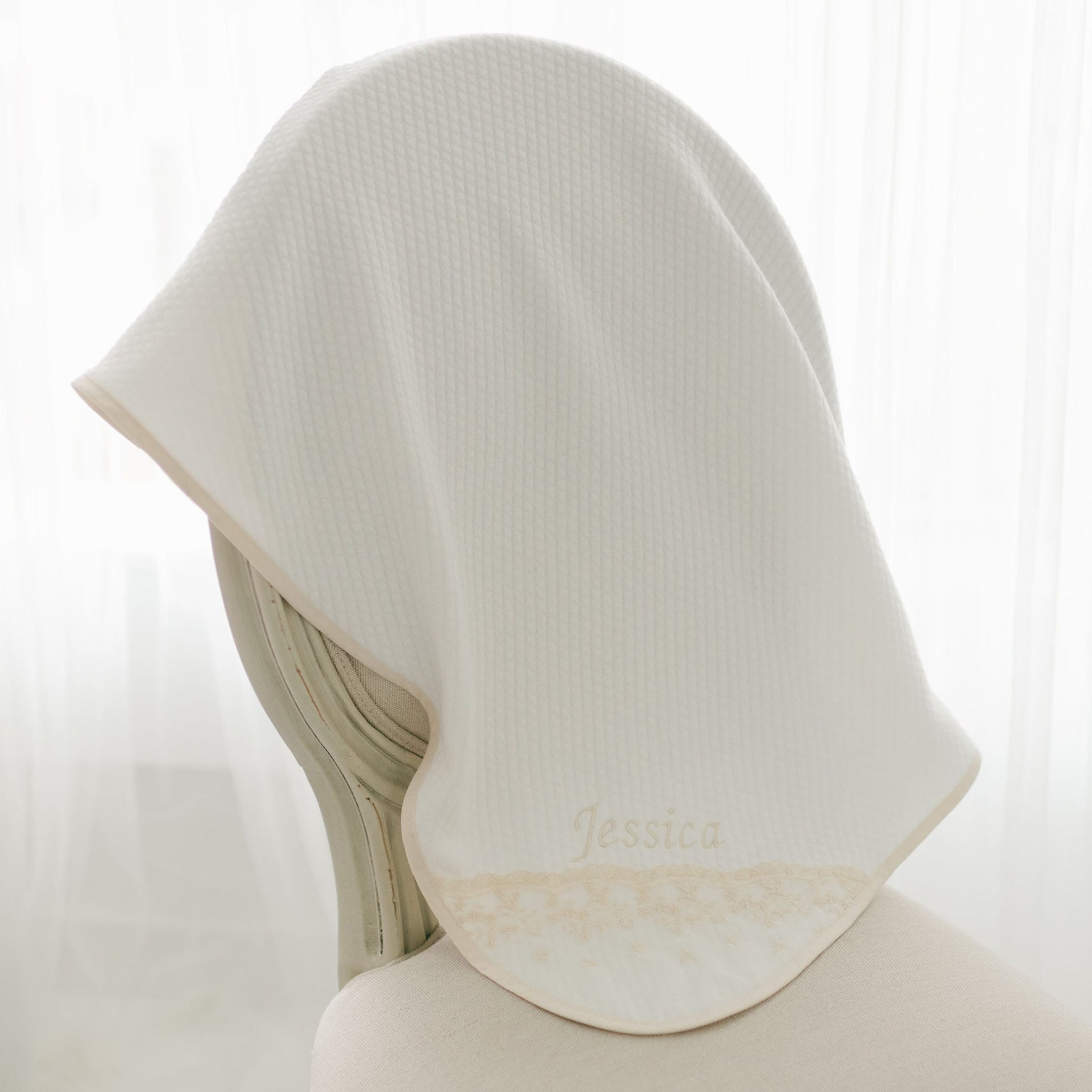 A white, handmade Jessica Personalized Blanket embroidered with the name "Jessica" is draped over the back of a chair. This Jessica Personalized Blanket has a textured surface and light brown trimming along the edge. The background is softly lit with white curtains, making it an elegant baby gift.