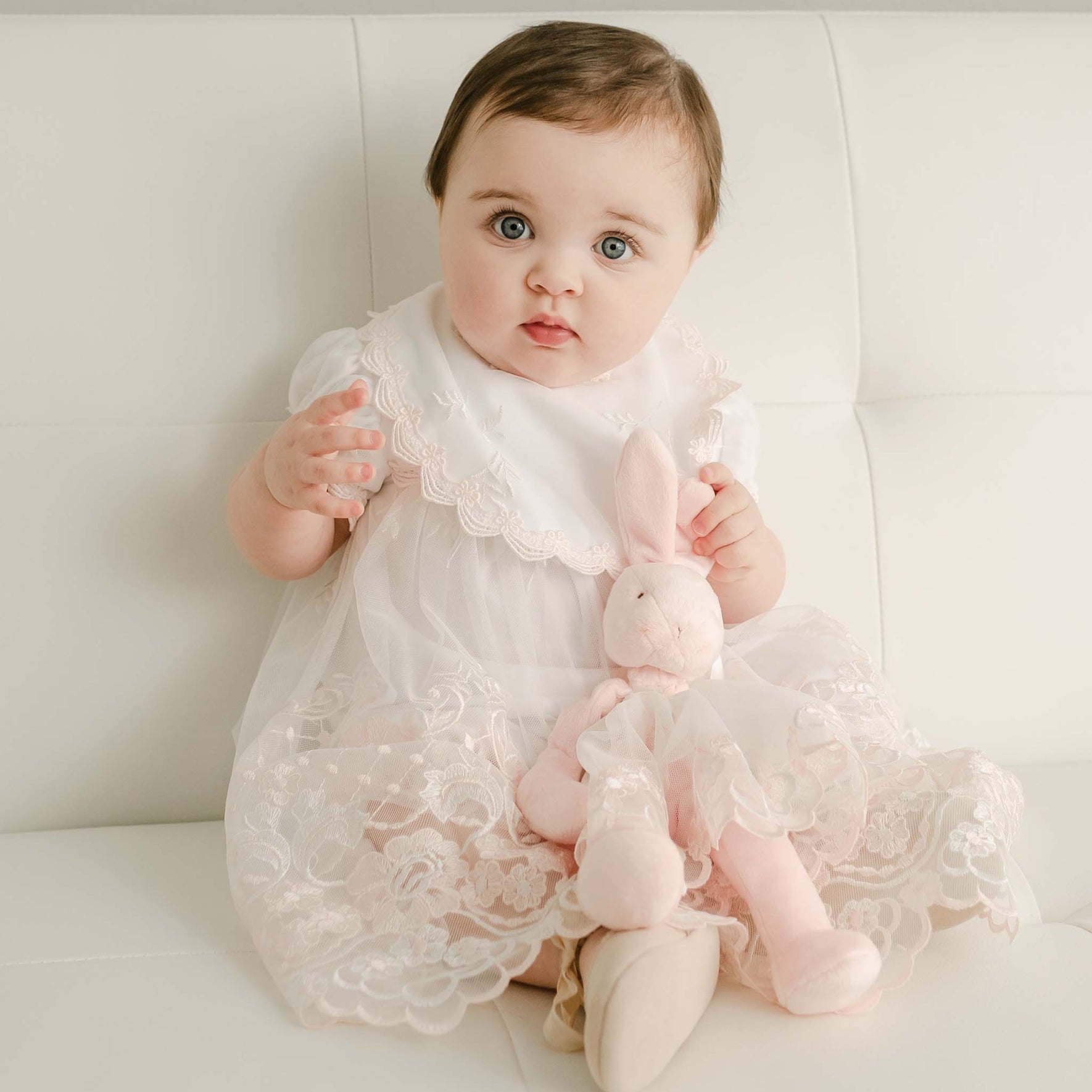 A baby wearing a Joli Bib Venice lace dress sits on a white couch, holding a pink stuffed bunny toy. The setting is bright and minimalist, emphasizing the delicate craftsmanship of the attire.