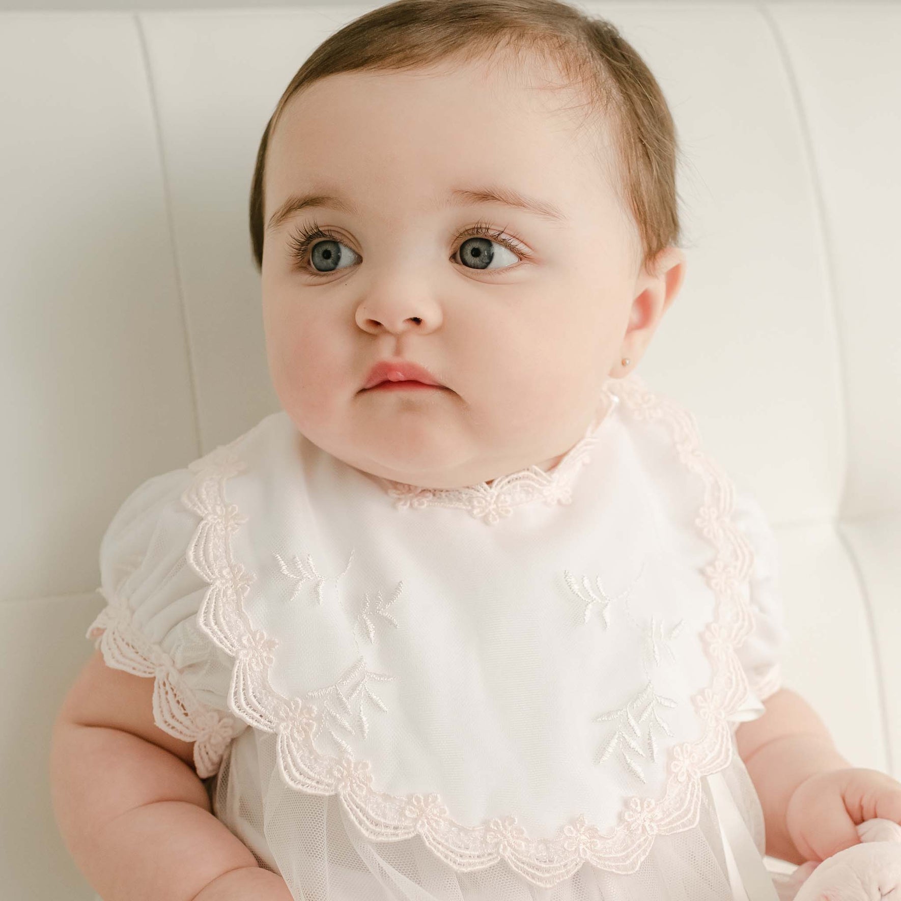 A baby with brown hair and large eyes is seated on a white surface, dressed in the Joli Bib christening romper. This exquisite outfit is embellished with elegant Venice lace and delicate embroidery, made from soft pima cotton that enhances its gentle charm. The baby gazes off to the side with a calm expression.