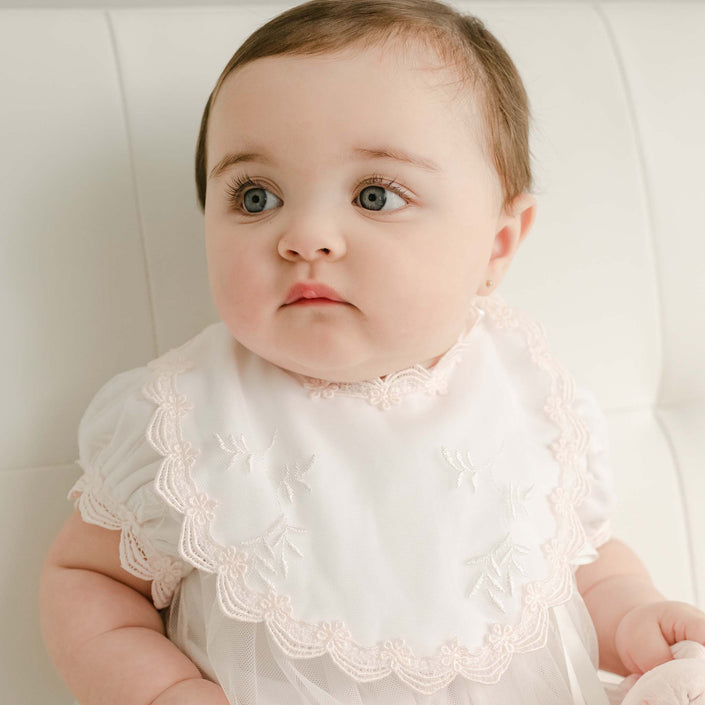 A baby with brown hair and large eyes is seated on a white surface, dressed in the Joli Bib christening romper. This exquisite outfit is embellished with elegant Venice lace and delicate embroidery, made from soft pima cotton that enhances its gentle charm. The baby gazes off to the side with a calm expression.