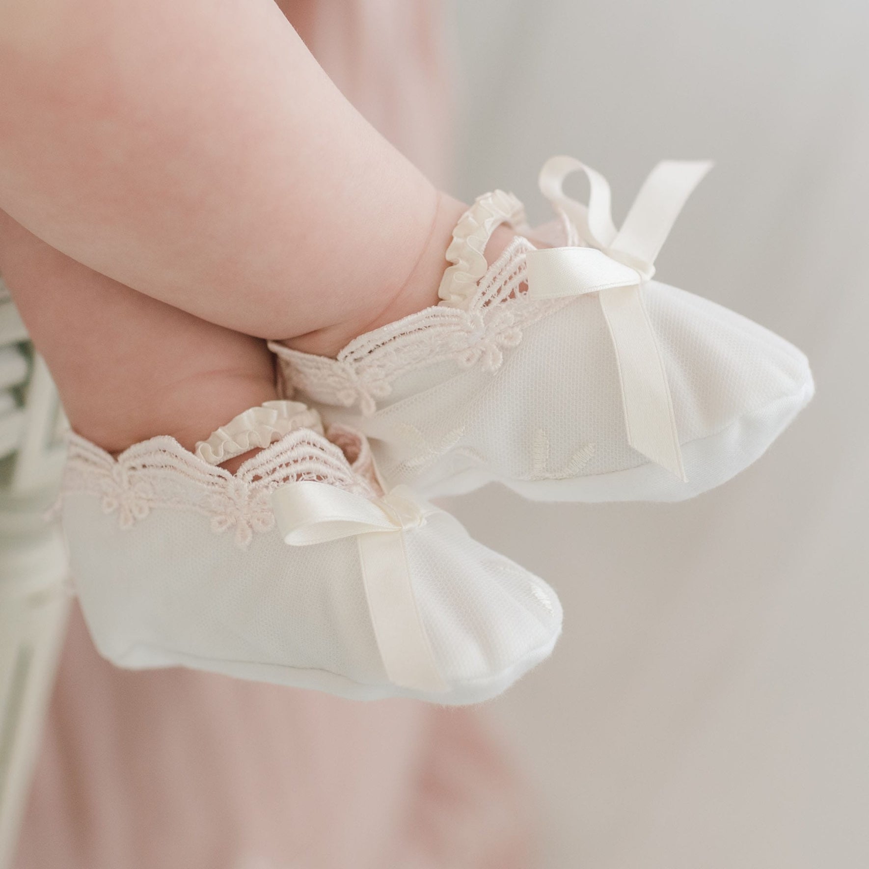 Close-up image of a baby's feet wearing white fabric shoes with floral lace trim and ribbon bows. The Joli Booties exude a delicate, vintage style, while the baby's legs are slightly blurred in the background.