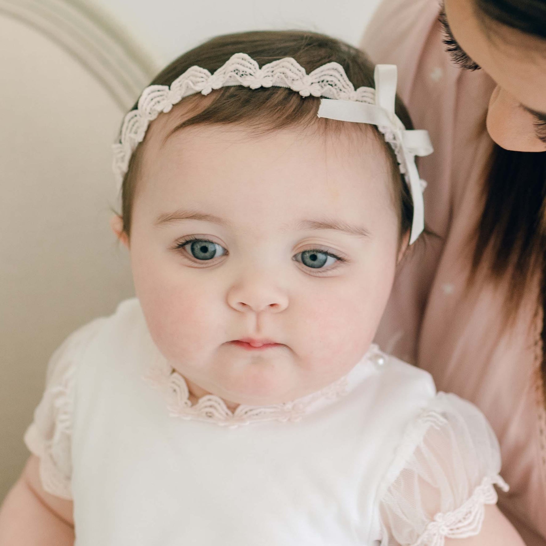 A baby dressed in white sits on an adult's lap, wearing a Joli Christening Headband featuring a delicate ivory silk bow. The adult, partially visible, gazes lovingly at the child against a neutral background.