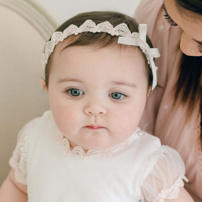A baby dressed in white sits on an adult's lap, wearing a Joli Christening Headband featuring a delicate ivory silk bow. The adult, partially visible, gazes lovingly at the child against a neutral background.