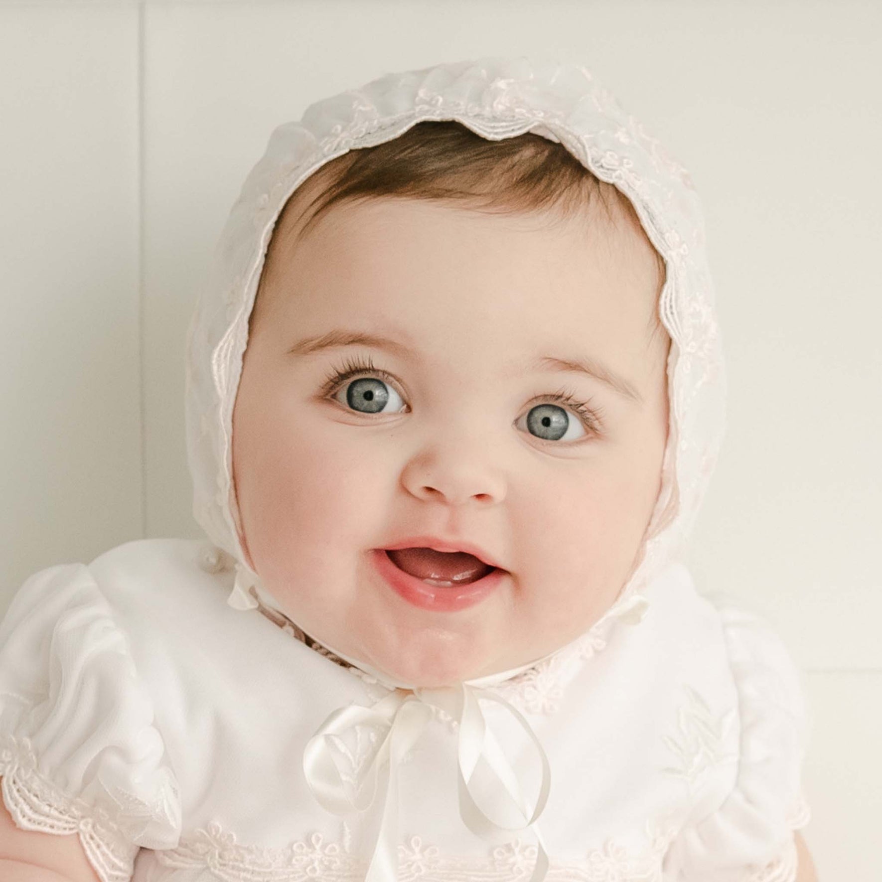 A baby girl in the Joli Fitted Christening Bonnet with lace detailing and matching baptism gown looks up with big blue eyes and a joyful expression. The bonnet's silk ribbon ties are cinched around her chin.
