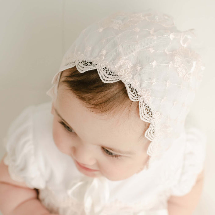 Top view of a baby girl wearing the Joli Fitted Christening Bonnet, featuring delicate floral lace and scalloped edges. The angle showcases the bonnet's embroidered netting over the pima cotton.
