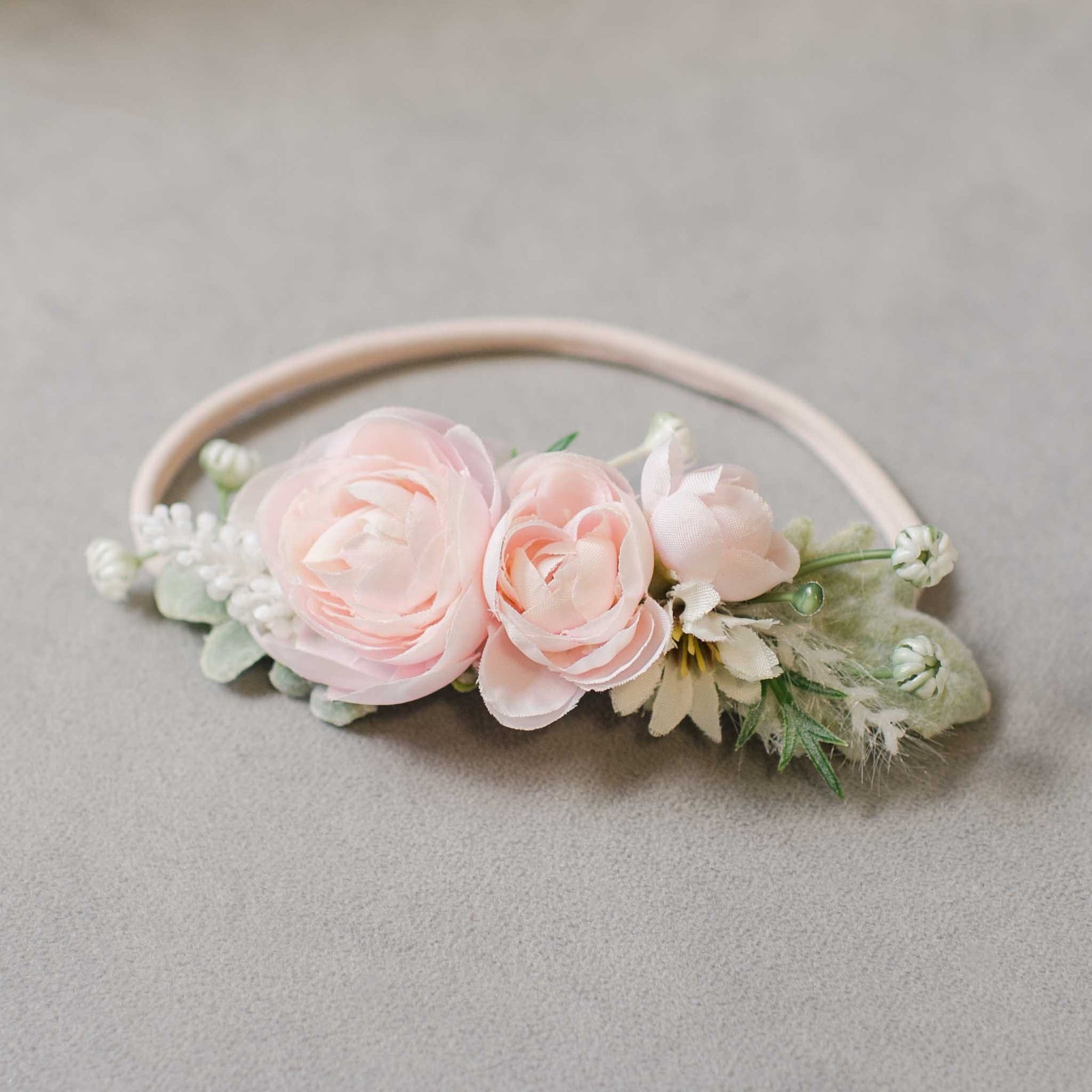 Introducing the Joli Flower Headband, a beautifully handcrafted accessory showcasing an exquisite arrangement of light pink roses, white buds, and lush green leaves, all artfully displayed on a sophisticated gray background.