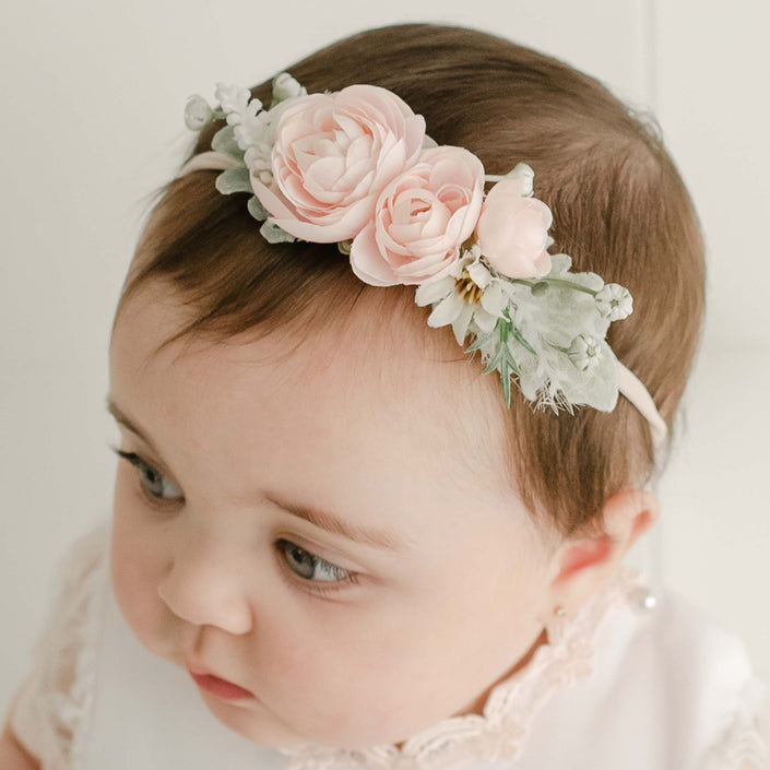 A baby wearing the Joli Flower Headband, embellished with pink roses and green leaves, gazes off to the side. The child is dressed in a light-colored outfit, with the delicate headband crafted from hand-stretched nylon.