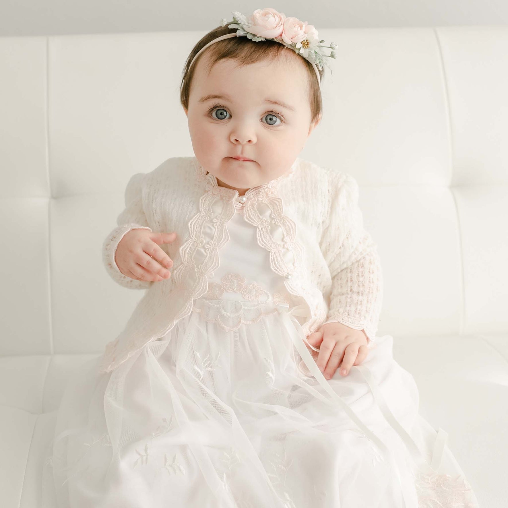 A baby sits on a white couch wearing the charming Joli Knit Sweater, beautifully adorned with Venice lace trim and delicate pearl-style buttons. Her look is completed with a floral headband featuring pink flowers, as she gazes directly at the camera.