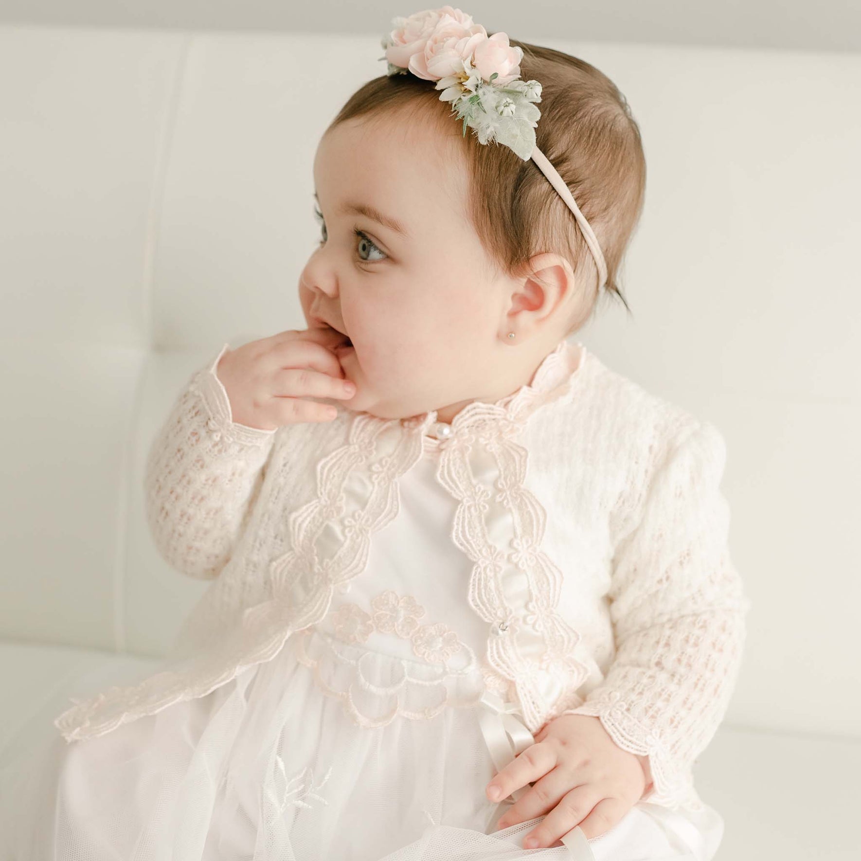 A baby girl wearing the Joli Knit Sweater in light pink, featuring delicate Venice lace trim and pearl-style buttons, sits on a white couch. She pairs it beautifully with a floral headband adorned with pink flowers, as she puts her fingers in her mouth and gazes to the side.