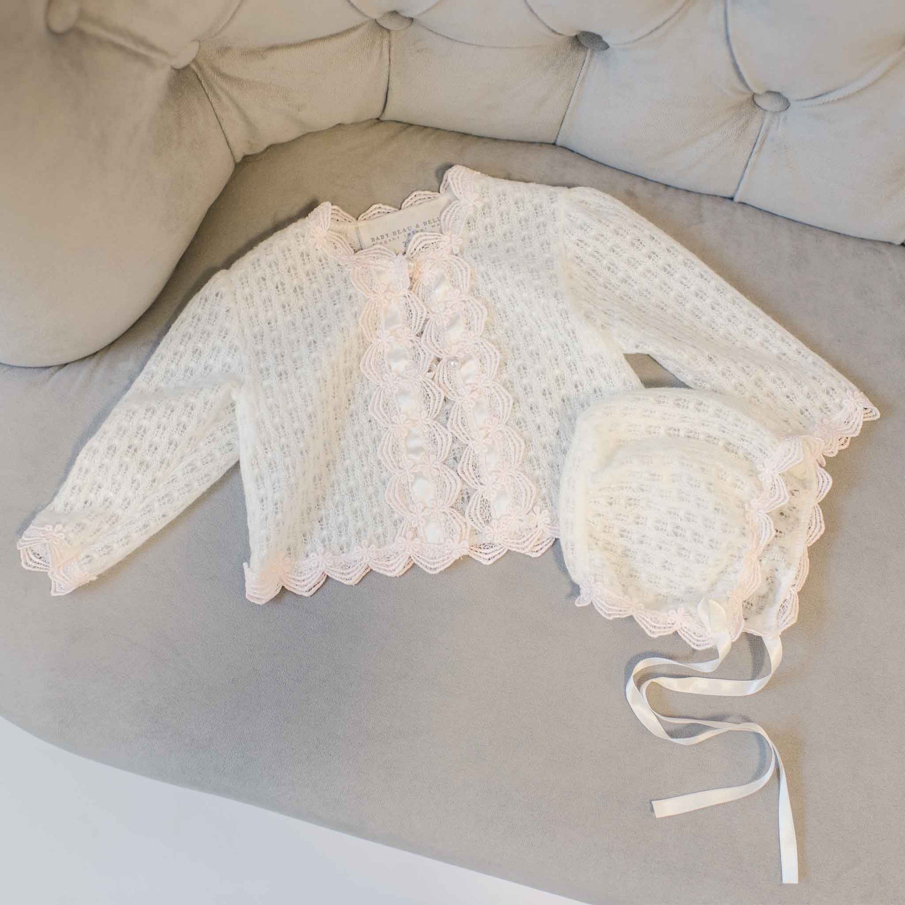 The Joli Knit Sweater & Bonnet set, displayed elegantly on a tufted gray cushion, showcases intricate lace patterns with Venice lace trim and delicate ribbon details. The ensemble is completed with pearl-style buttons for an extra touch of charm.