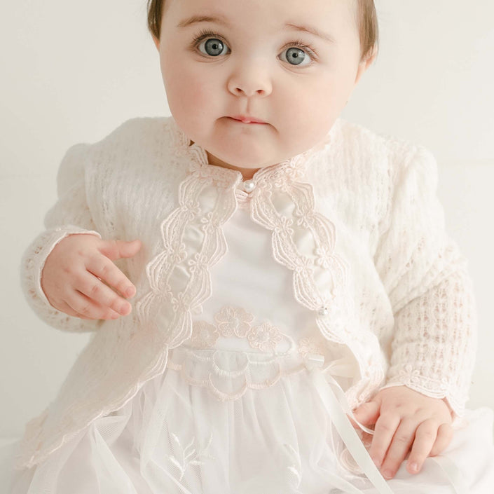 The baby, with light eyes and short dark hair, wears a delicate white embroidered dress featuring Venice lace trim. Her outfit is completed by the Joli Knit Sweater & Bonnet set, which is adorned with pearl-style buttons that enhance her look as she sits against a light background, gazing at the camera with a neutral expression.