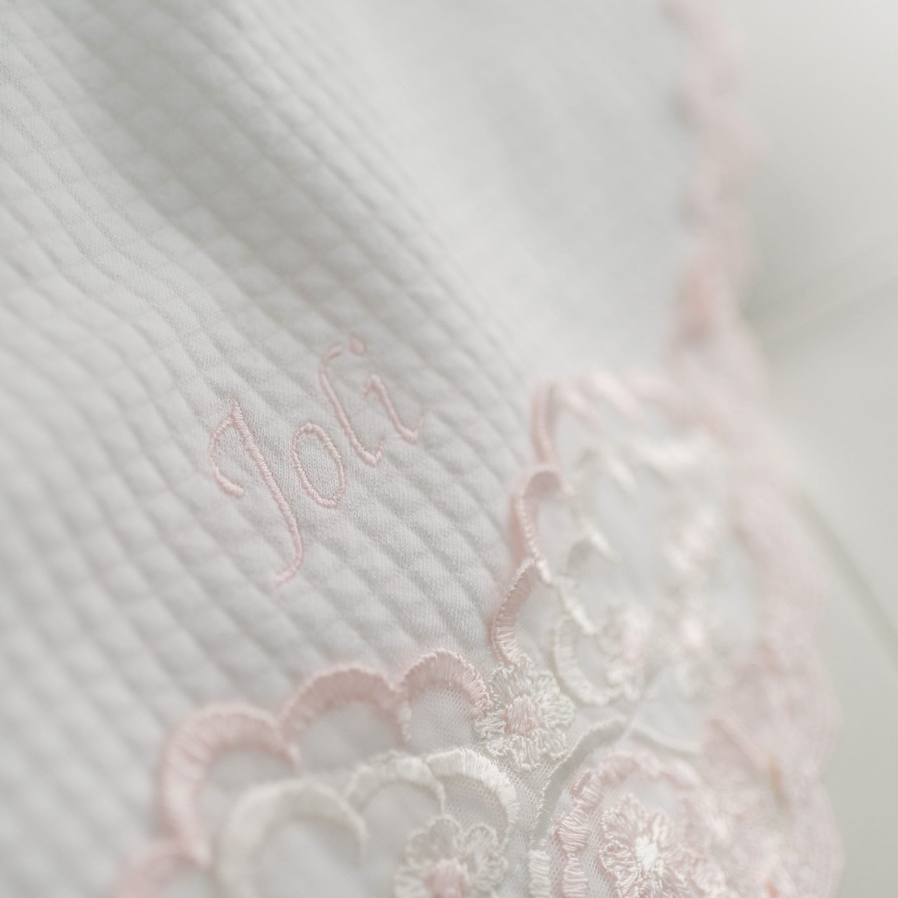 A close-up photo of a Joli Personalized Blanket with the embroidered name "Joli" in pink thread, partially overlaying an intricate white and pink lace design.