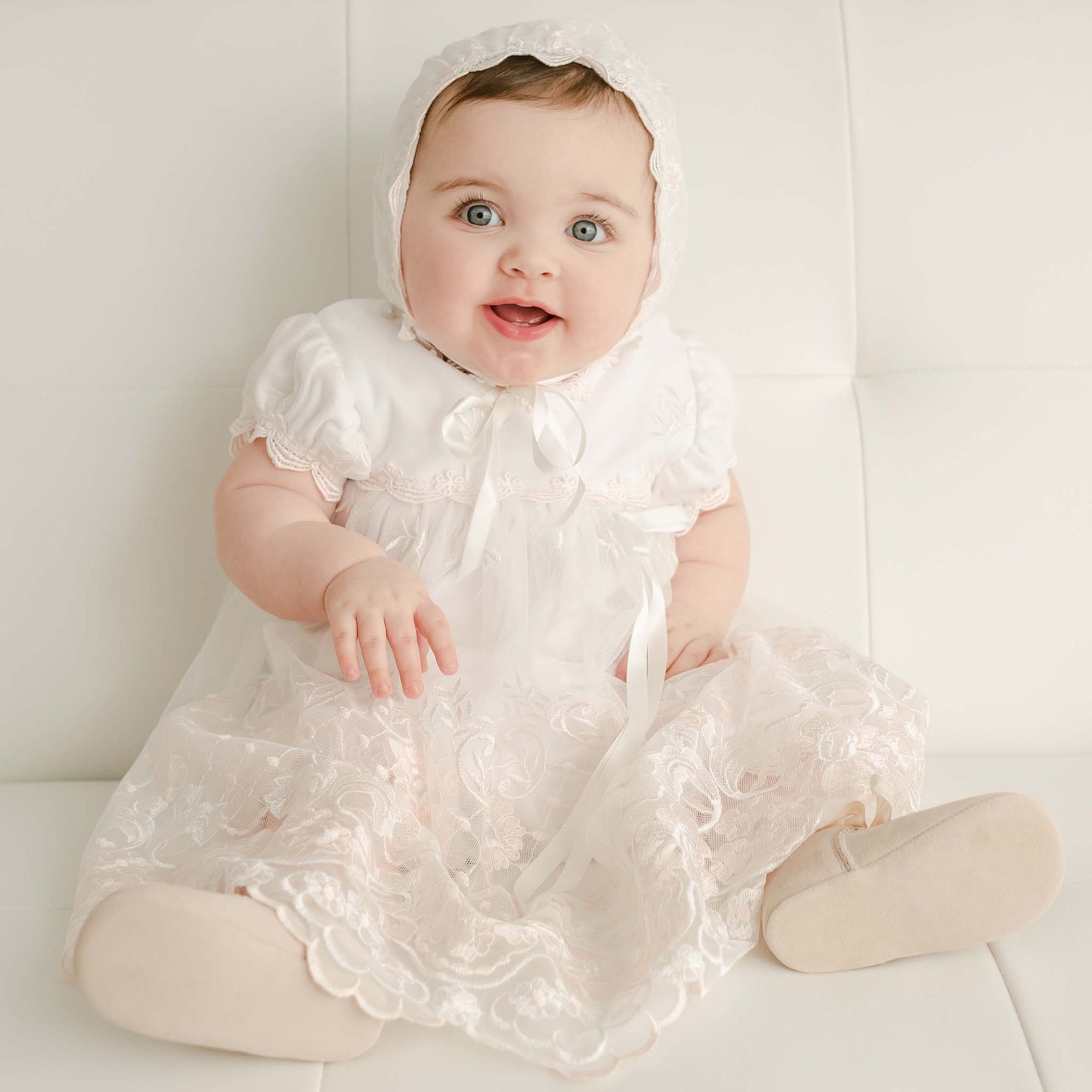 A baby dressed in the Joli Romper Dress, featuring intricate lace patterns and ribbon details, sits on a white surface. The cheerful baby captures hearts with wide eyes and a slight smile, wearing the delicate embroidered netting lace ensemble complete with a bonnet.