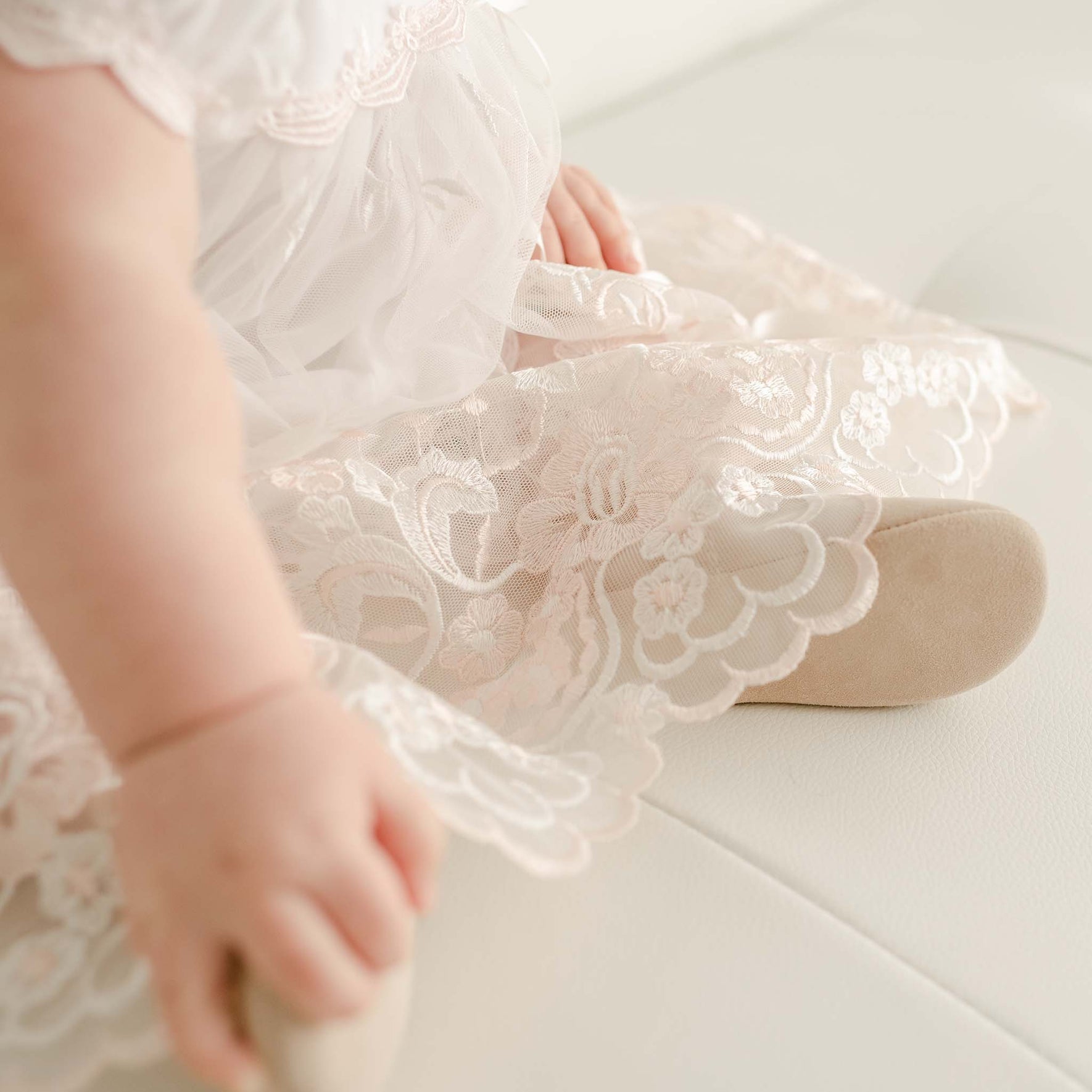A close-up of a baby sitting on a white surface, wearing the Joli Romper Dress, an embroidered netting lace outfit adorned with floral patterns. The baby's hand rests gently on the dress, and their feet are cozily covered with soft beige shoes.