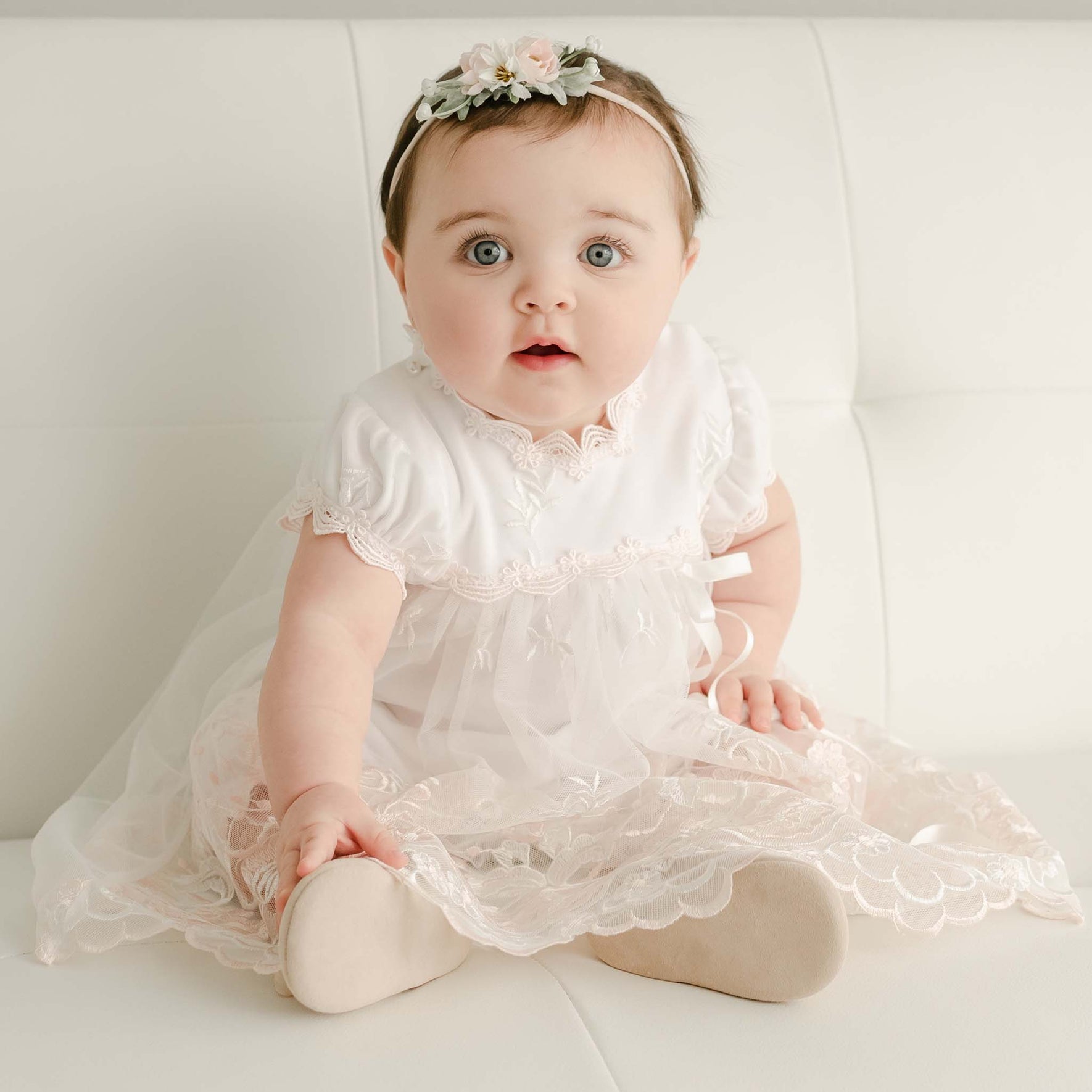 A baby in a Joli Romper Dress with embroidered netting lace sits on a white sofa. Wide-eyed and adorned with a floral headband, the little one looks charming. The delicate skirt and sleeves feature sheer, lace-like fabric, while the baby remains barefoot in this adorable scene.