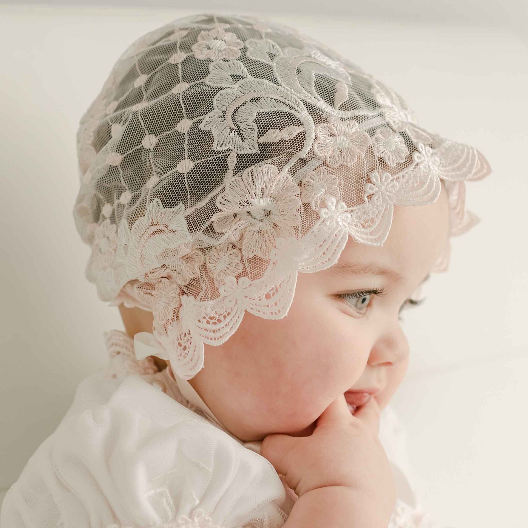 Baby girl in the Jolie Sheer Christening Bonnet and matching dress looks away from the camera with one hand in her mouth. The side view highlights the intricate embroidered netting of the bonnet.