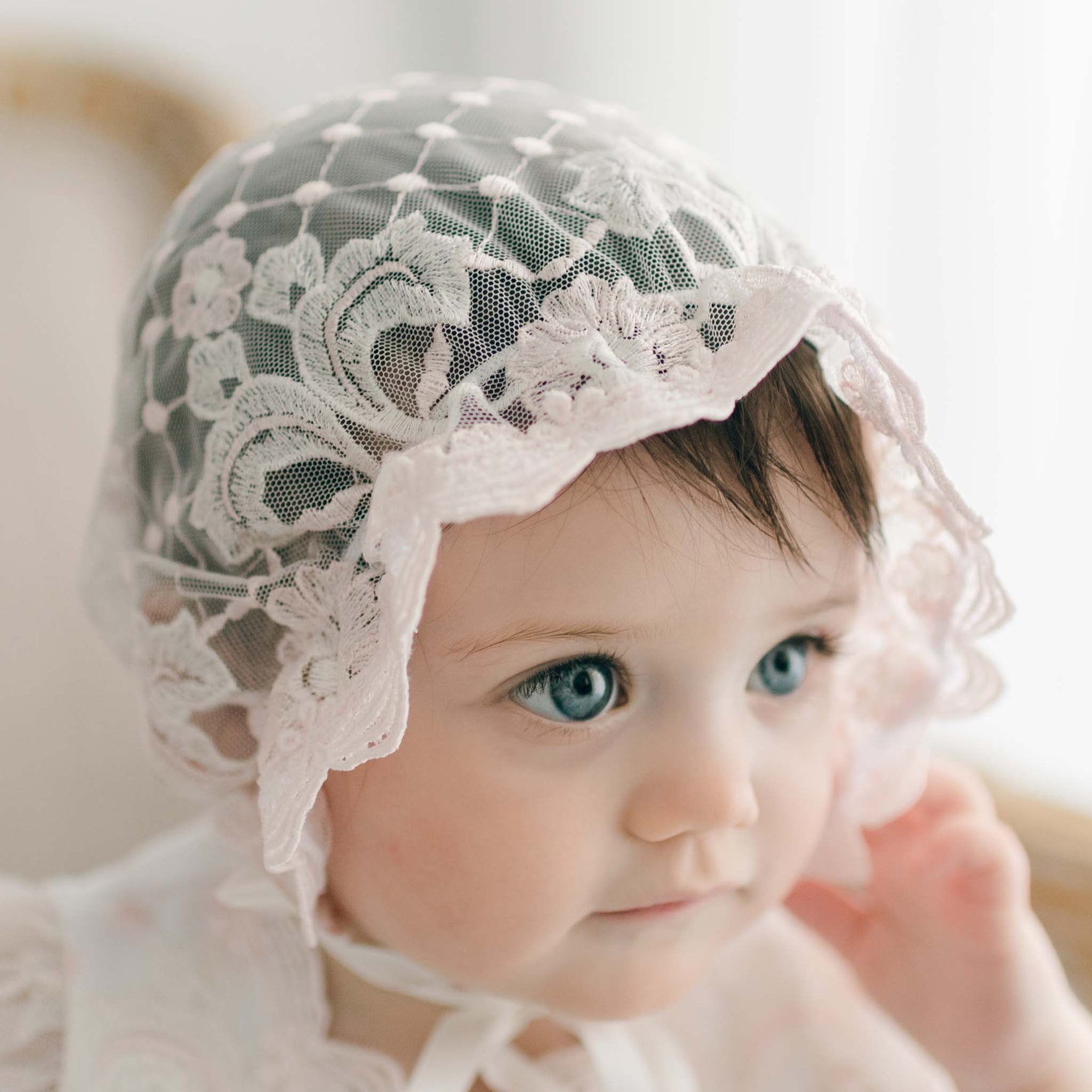 Close-up of a baby girl in the Jolie Sheer Christening Bonnet, featuring intricate floral lace embroidery and ruffled edges. The bonnet is tied with silk ribbons, framing her face as she looks off to the side.