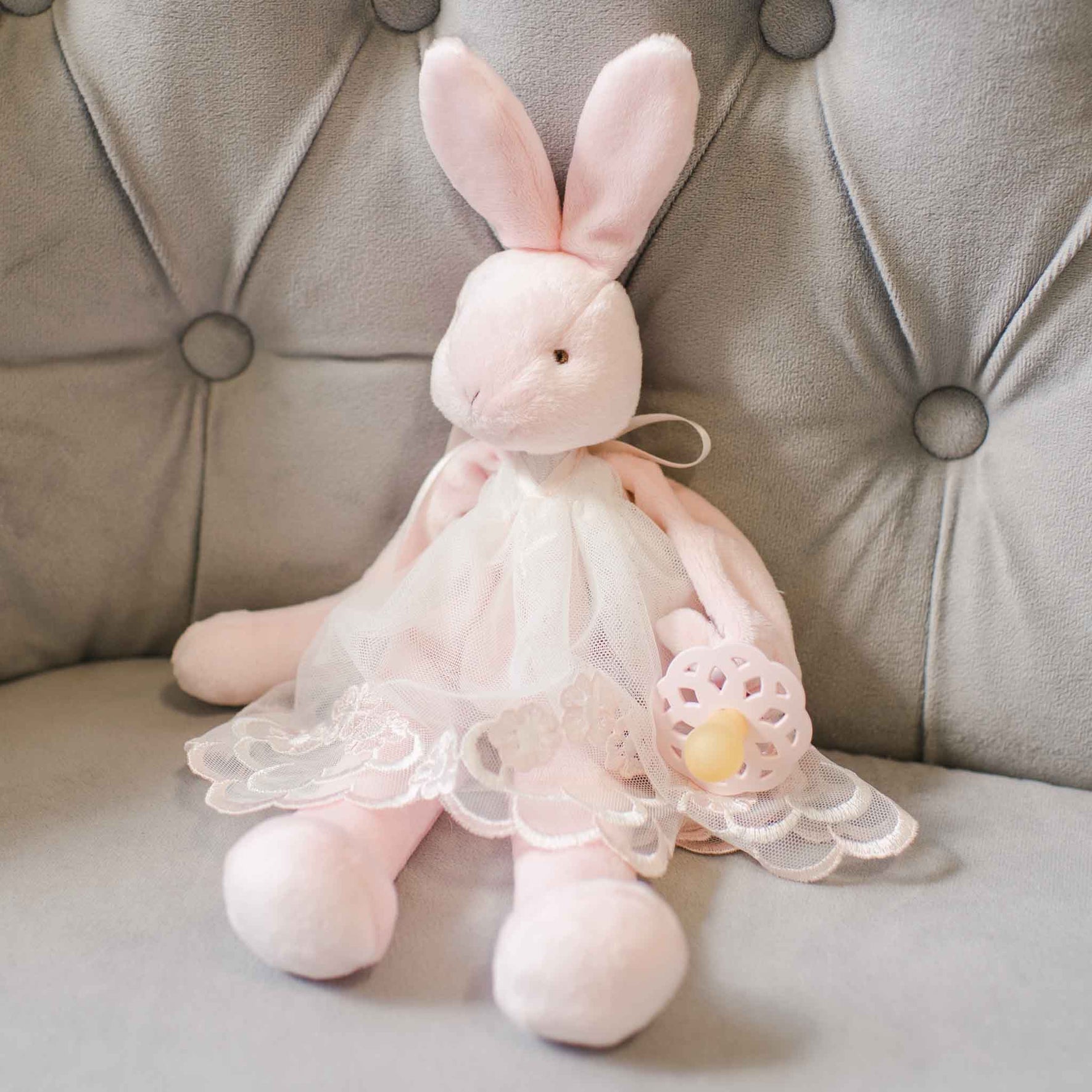 A Joli Silly Bunny Buddy | Pacifier Holder, baby safe and softly adorable, sits on a tufted gray chair. Dressed in a white lace dress, this plush pink bunny holds a pink and yellow teething ring and can easily velcro onto a stroller for gentle travels together.