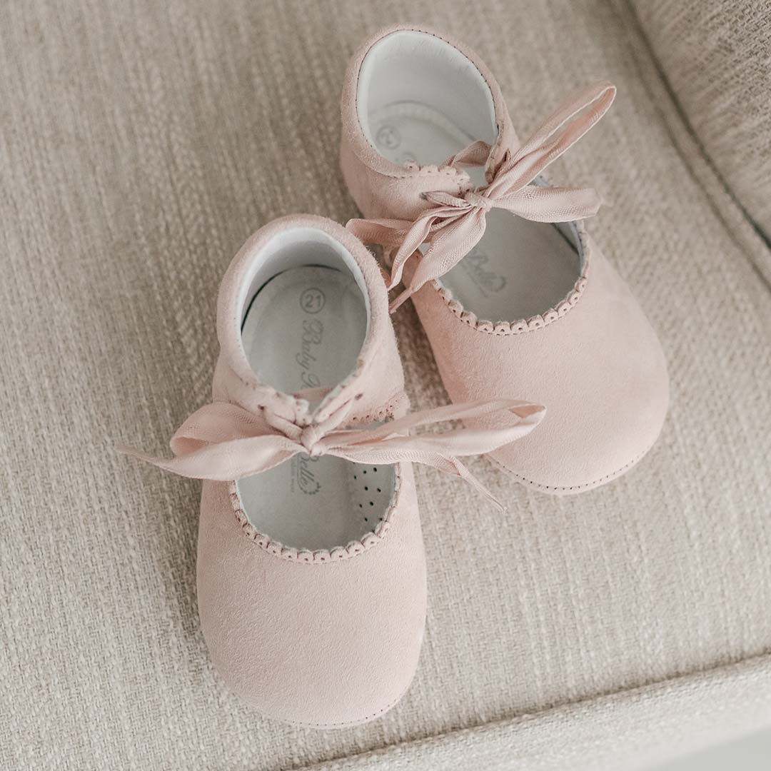 A pair of pink handmade baby shoes are placed on a beige textured surface. The Joli Suede Tie Mary Janes, crafted by Baby Beau & Belle, have a soft, suede-like material with bows tied at the top and scalloped edges around the opening.